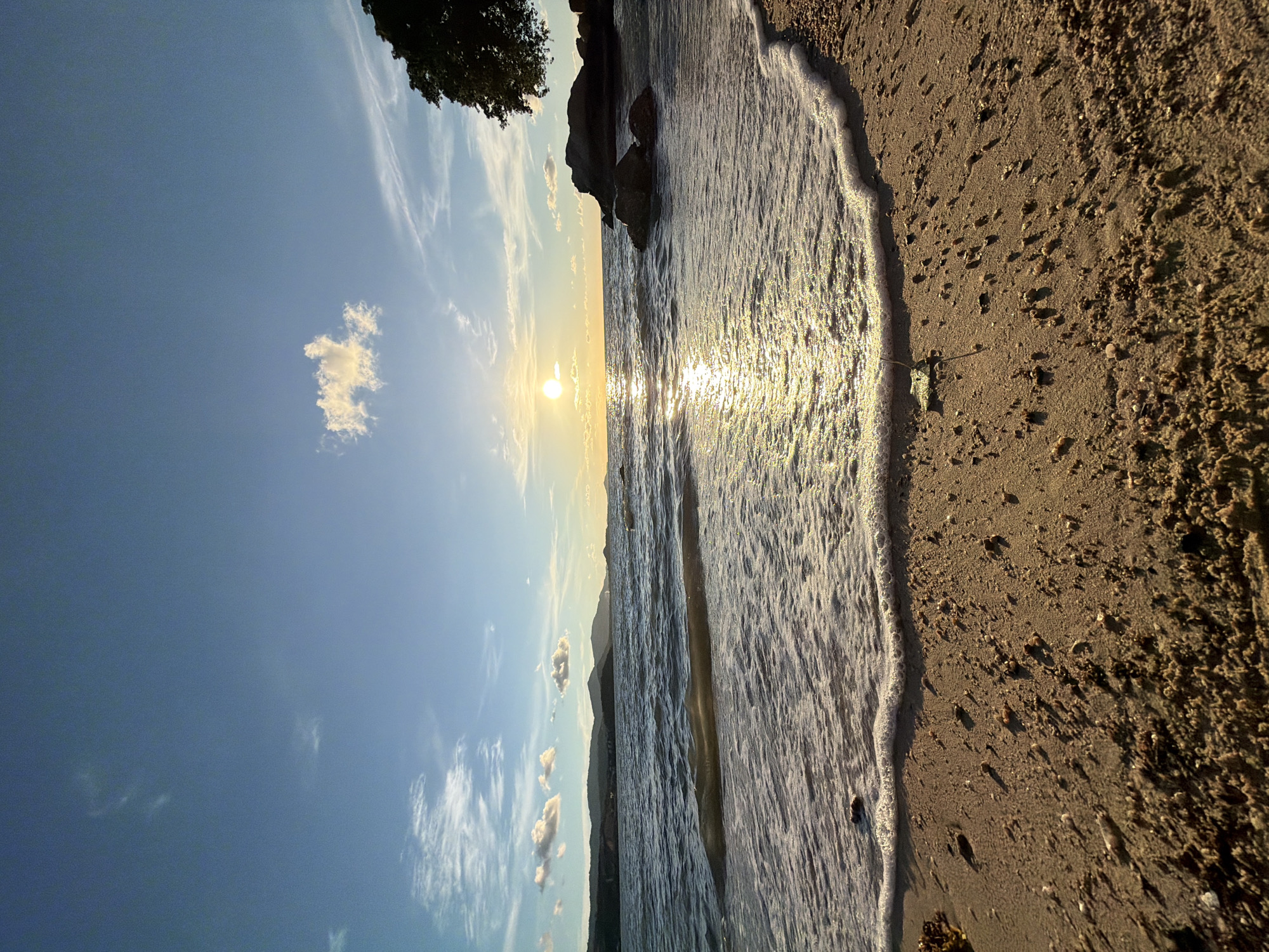 The media file depicts a serene beach scene at sunset in Baan Bo Phut, Thailand. The sun, a brilliant orb, hangs low on the horizon, casting a warm, golden glow across the water and sand. Its reflection shimmers on the gentle waves as they lap against the shore.
The foreground is dominated by the sandy beach, textured with small pebbles and scattered debris. The water's edge is marked by a line of frothy white foam, indicating the tide's ebb and flow. In the distance, a faint silhouette of hills or mountains can be seen against the vibrant sky.
The sky itself is a canvas of blues and soft whites, with wispy clouds streaking across the expanse. A single, fluffy cloud is particularly noticeable, catching the sunlight. The dark silhouette of a tree branch or foliage frames the upper left corner of the image, adding depth and a touch of natural drama.
The overall impression is one of tranquility and natural beauty. The warm light of the setting sun evokes a sense of peace and calm, characteristic of a perfect beach sunset. There is no visible text in the media file. The scene suggests a quiet moment of appreciation for the natural world, possibly at the end of a day in this picturesque Thai location.
