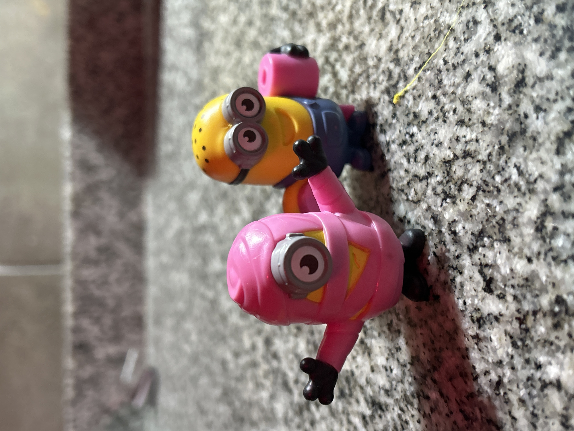 Two Minion toys are positioned on a speckled grey and white surface. The top minion is yellow with two eyes, wearing grey goggles, a blue outfit, and a pink head accessory. The bottom minion is pink with a single eye wearing a grey goggle. A thin yellow strand rests on the surface near the top minion. The background shows an out-of-focus grey vertical surface and an adjacent slightly blurred grey and white surface. The location is Buenos Aires, Argentina.