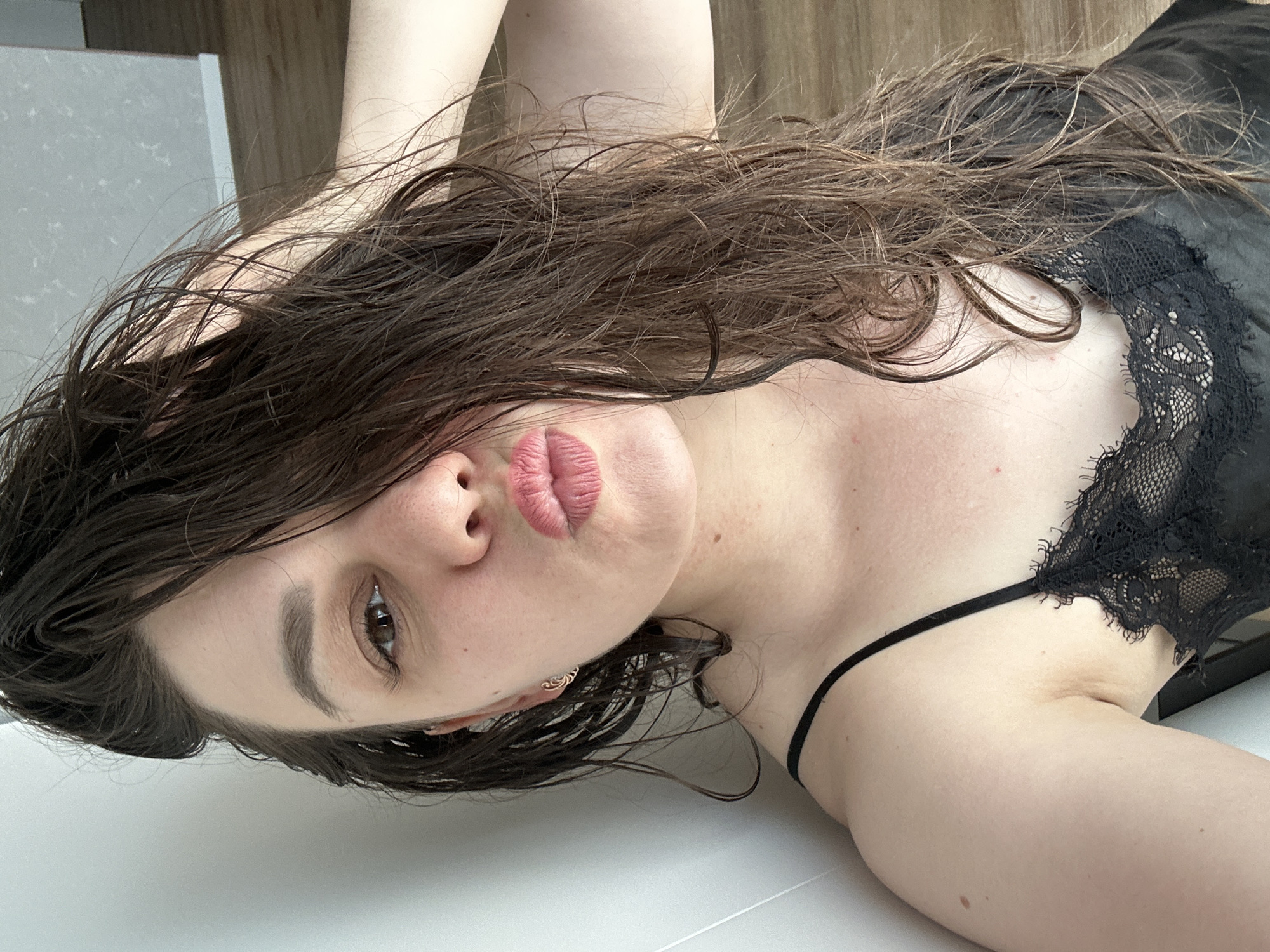 A woman with wet, long brown hair is lying on a white surface, possibly a counter or table. She is wearing a black lace-trimmed garment with thin straps. Her face is in focus, revealing fair skin, brown eyes, and pink lips. She is wearing a small, ornate earring on one side. The background shows a glimpse of a white wall or surface and a brown wooden surface. There is no information in the image to indicate that this is in Chaikovsky, Russia.