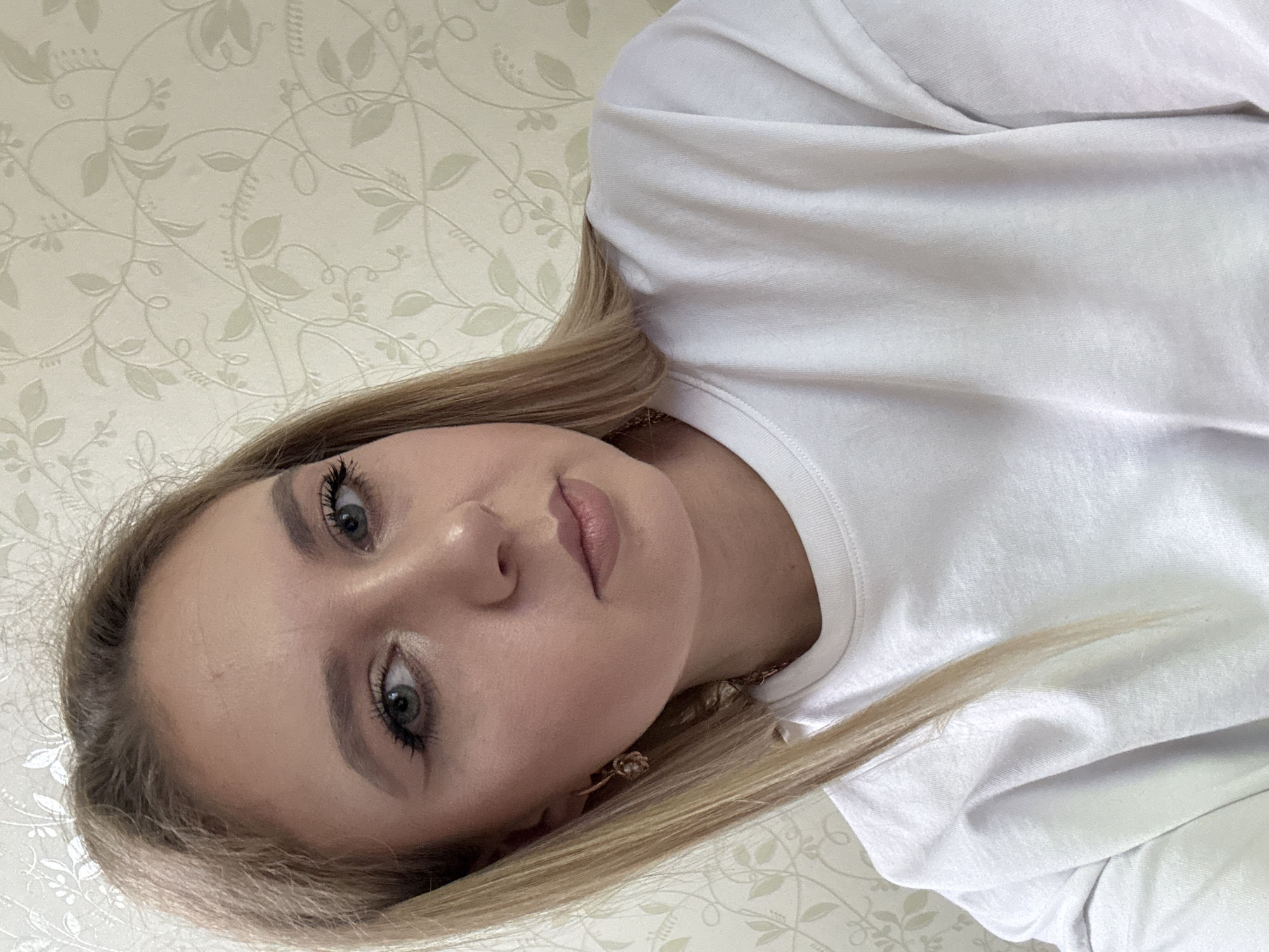 A young woman with blonde hair, wearing a white t-shirt and a rose gold earring, poses for a selfie in front of a floral patterned wallpaper. She has light makeup on, including eyeliner and mascara. Her hair is straight and parted to the side, falling over her left shoulder. The background appears to be a domestic interior, likely a room in a house or apartment. The lighting suggests it's daytime, possibly indoors with natural light.