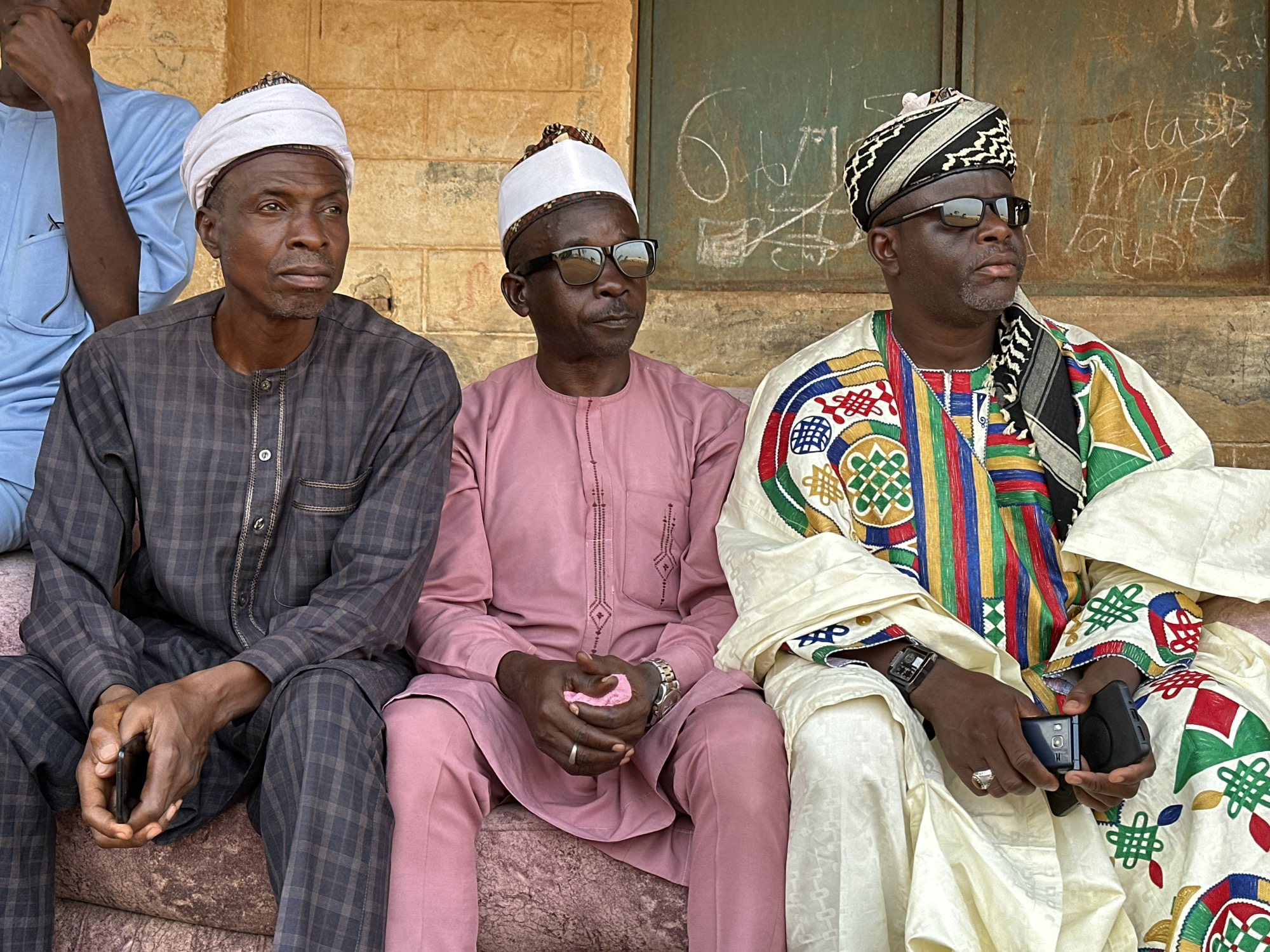 Three men sit side-by-side outdoors. The man on the left wears a gray plaid robe and a white turban. The man in the center wears a pink suit and white cap and is wearing sunglasses. The man on the right wears a white patterned robe, a striped and patterned turban, and sunglasses. He is holding a small black device. A fourth man, in a blue shirt, is partially visible at the left edge of the frame. The background is a beige wall with green graffiti. The location appears to be a public space. The city is Unknown, Unknown.