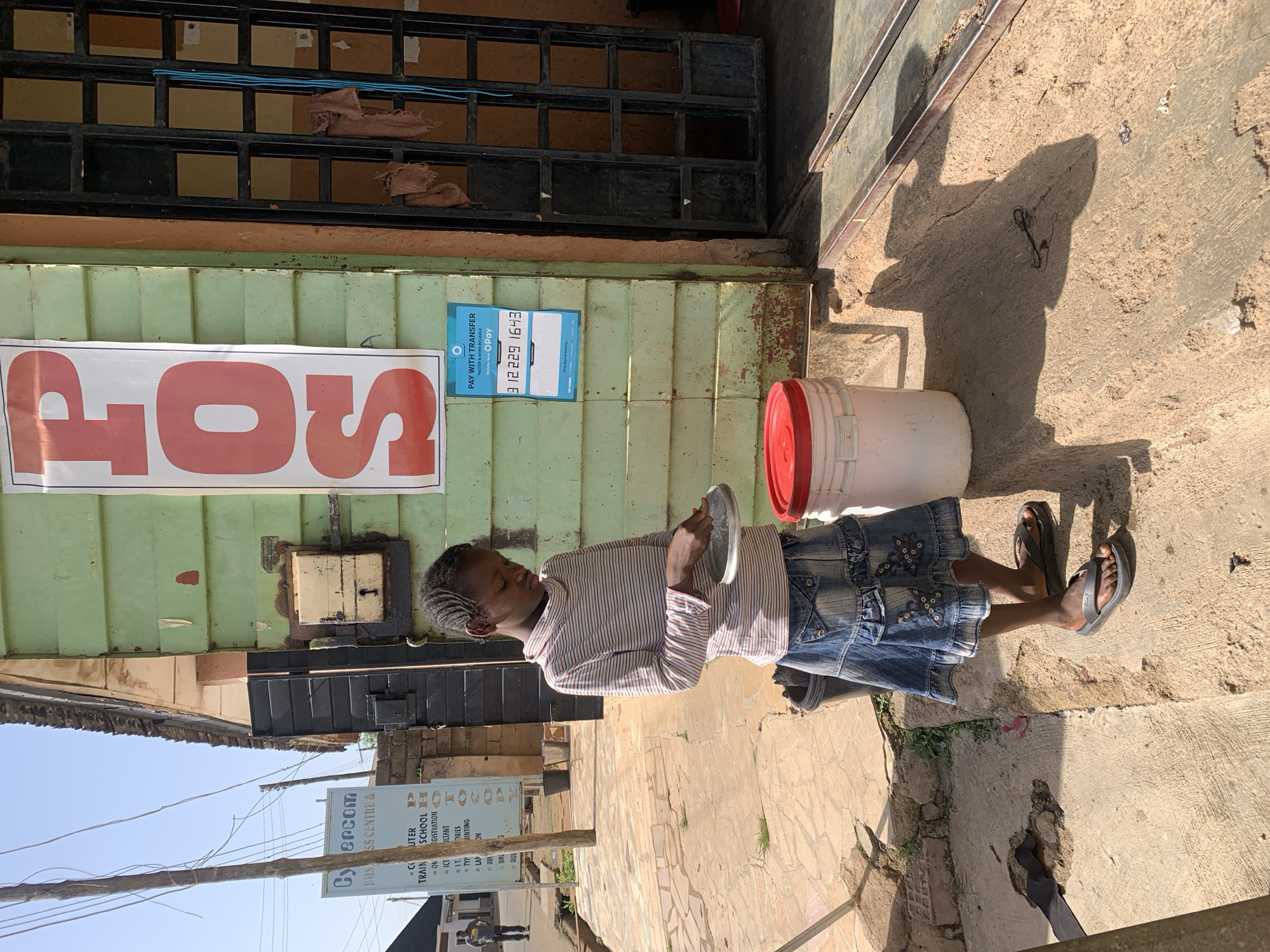 An individual stands outside a business in Jos, Nigeria, holding a circular metal object. The business appears to be a Cybercom business center and computer school, based on a sign displayed. The individual is wearing a striped shirt and denim skirt. Behind them is a green door with the word "POS" written on a sign. A white bucket with a red lid is positioned nearby.