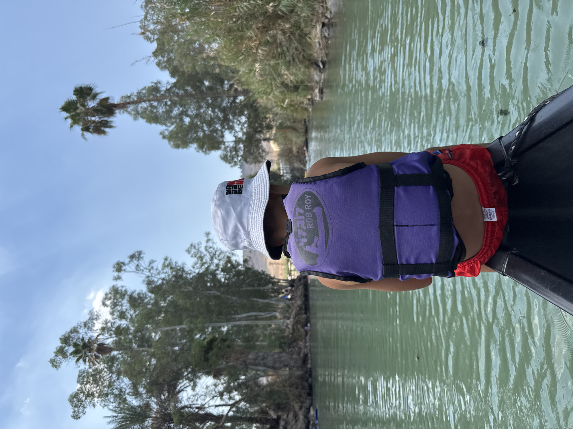 In Emek HaYarden Regional Council, Israel, a young person wearing a white bucket hat and a purple life vest with "ROB ROY" visible on it sits facing away from the camera, in what appears to be a kayak or boat. Red swimming shorts are visible below the life vest. The person is on a body of water with green, rippling water. The surrounding environment is lush with trees, including palm trees, and dense vegetation along the banks of the water. The sky above is blue with scattered clouds. In the background, other people can be seen in the water and along the shore.
