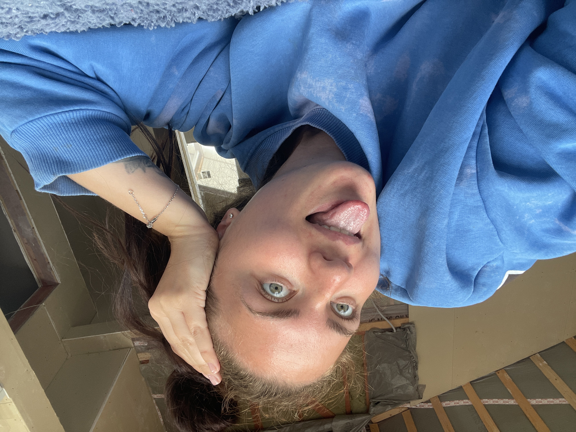 A young woman is pictured in a selfie-style shot. She is wearing a blue hooded sweatshirt and has her hair pulled up in a bun. She is sticking her tongue out and has a playful expression on her face, with her eyes wide open and looking at the camera. Her right hand is resting on her head, and she is wearing a silver bracelet on her left wrist. The background appears to be an unfinished room with wooden beams and drywall. The lighting suggests it may be daytime, with natural light entering the space. The overall impression is informal and candid.