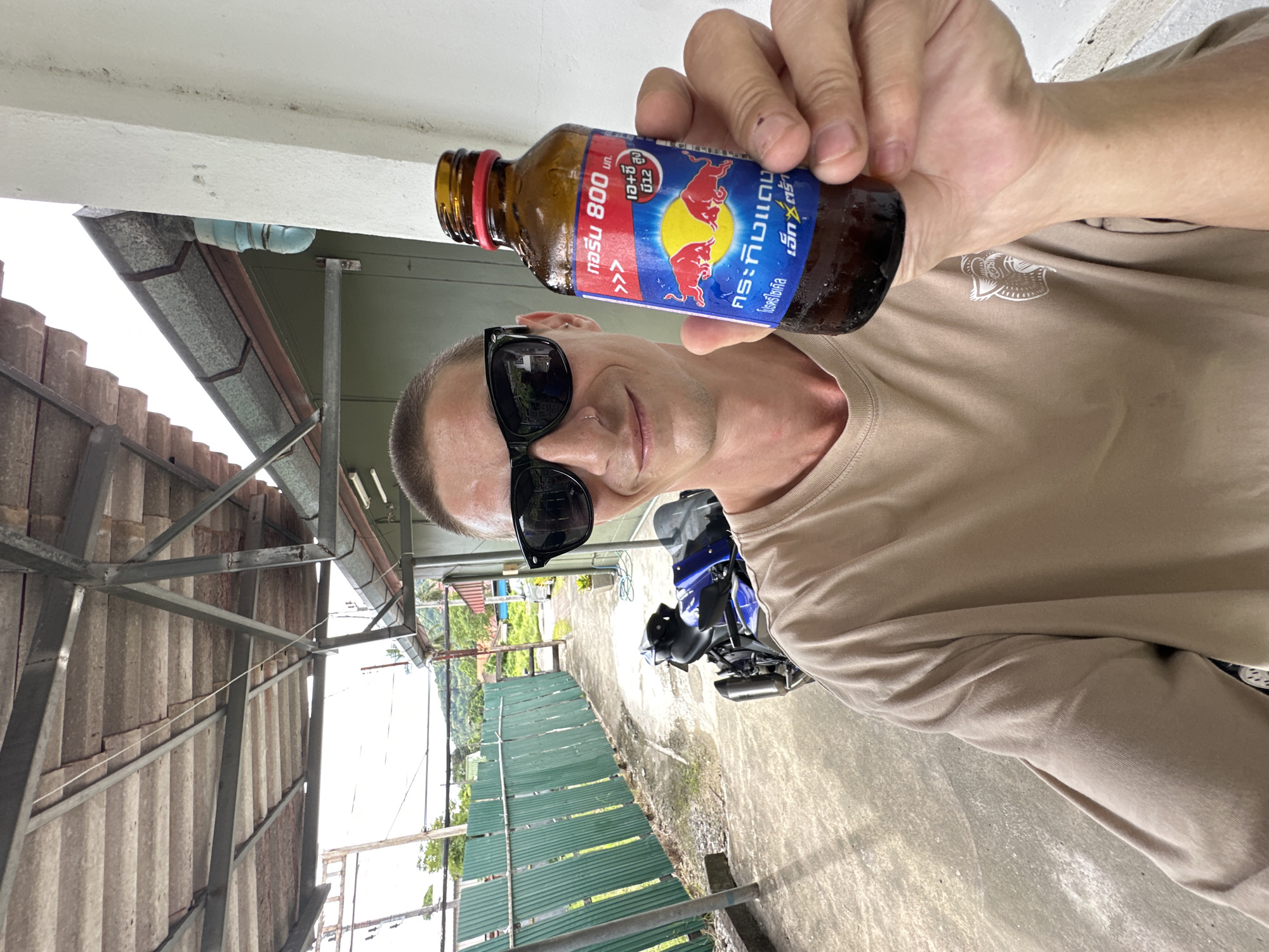 A person is holding up a small bottle of Red Bull in their right hand, showing the label to the camera. The bottle is brown glass with a blue and yellow label featuring the iconic Red Bull logo of two bulls charging. The label has text in Thai, which appears to be related to the drink's ingredients and name. The person is wearing sunglasses and a tan t-shirt with a subtle graphic on it. They have a nose ring and short hair.
The background suggests an outdoor setting, possibly a sheltered area like a carport or under a roof. There's a metal structure supporting a corrugated roof on the left. A green corrugated metal fence runs along the right side of the background. A blue motorcycle is parked in the mid-ground. Beyond the fence, there are hints of lush green vegetation and a hillside, suggesting a tropical or sub-tropical environment. The lighting appears to be natural daylight, indicating it is daytime, and the overall atmosphere seems casual and relaxed. The presence of a motorcycle and the style of the buildings hint at a location in Southeast Asia, consistent with the specified location of Karon, Thailand. The image is a selfie, with the person taking the photo themselves.