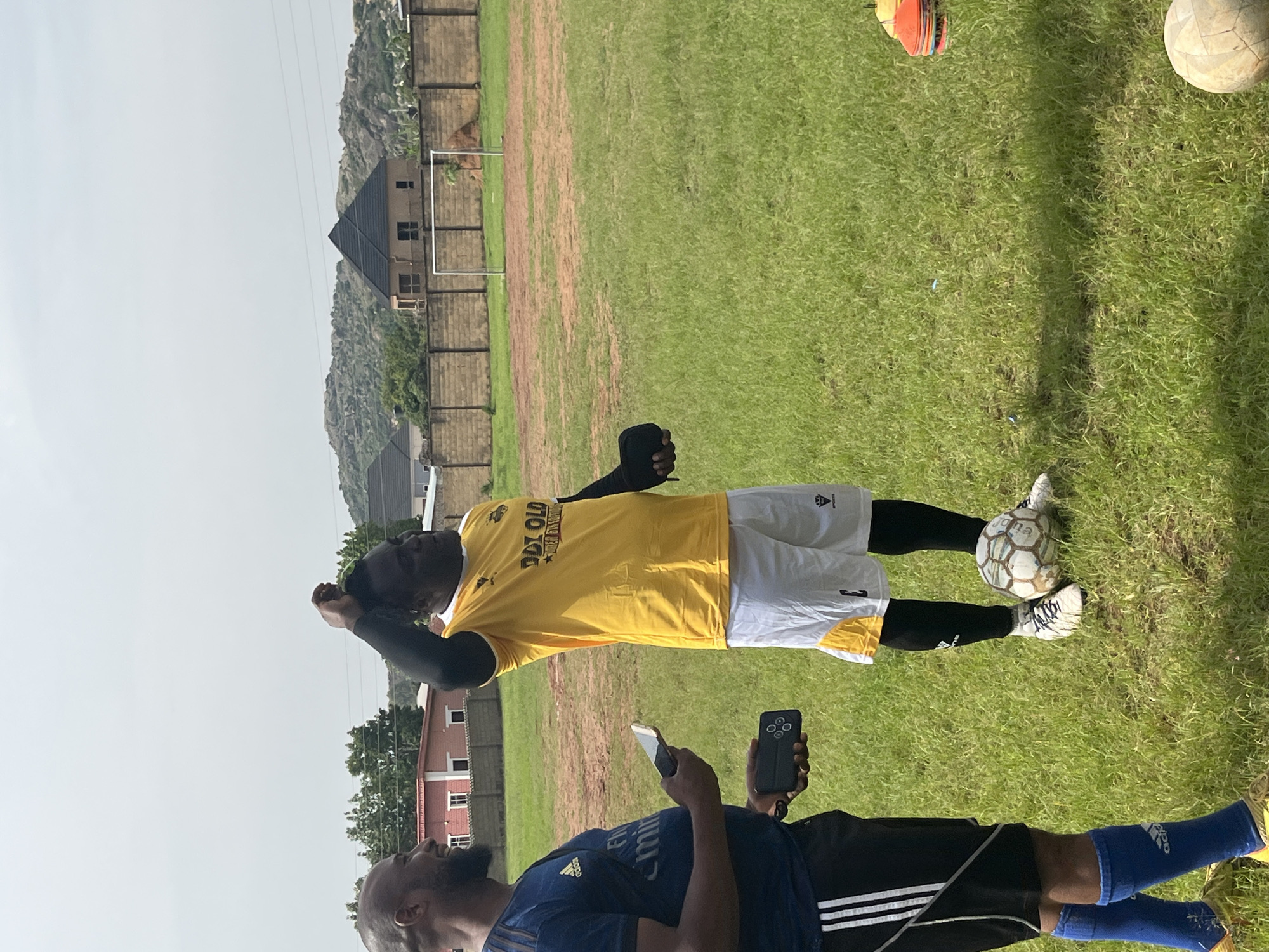 Two men stand on a grassy field, presumably in Jos, Nigeria, with buildings and rocky hills in the background. One man, on the left, wears a dark blue Adidas shirt and black shorts with white stripes, holding a smartphone and a small black device. The other man, on the right, wears a yellow shirt with text that appears to read "OLD" and white shorts with yellow accents. He is wearing black socks and white soccer cleats, with a soccer ball at his feet. He has his hand on his head. The field is green and appears to be a soccer field, with a goalpost visible in the background. The sky is overcast.