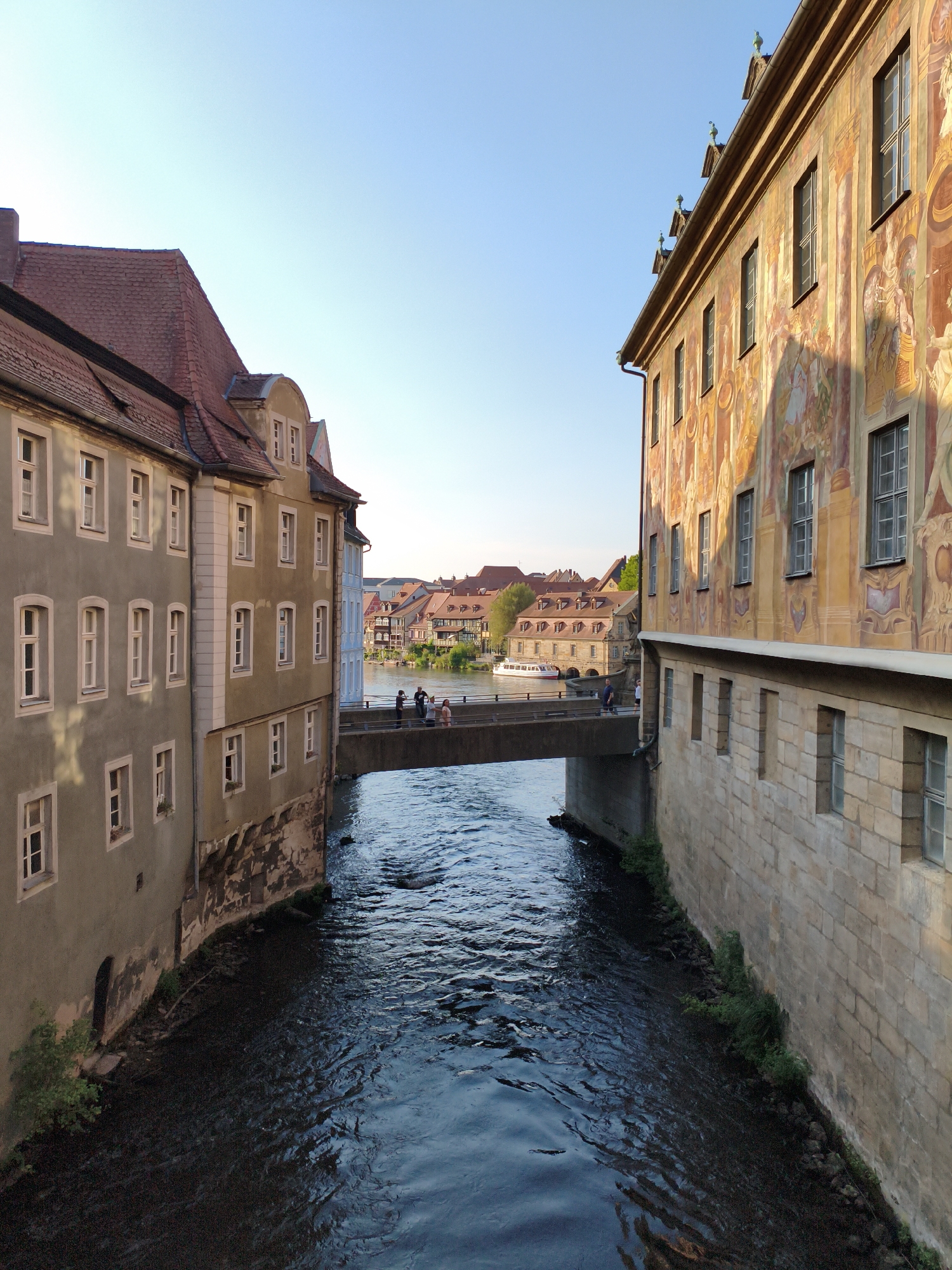 The image shows the Regnitz River in Bamberg, Germany, flowing between historic buildings. On the left, a multi-story building with a red tiled roof and numerous windows lines the riverbank. On the right, a larger, ornate building features a vibrant mural depicting figures and scenes, with a stone foundation at the river level. A pedestrian bridge spans the river, connecting the two sides. Several people are visible on the bridge, walking and conversing. In the background, more buildings and trees are visible, with a clear blue sky overhead. The water in the river is dark and reflects the light, with visible ripples and currents. The overall scene captures a picturesque view of Bamberg's riverside architecture and atmosphere.