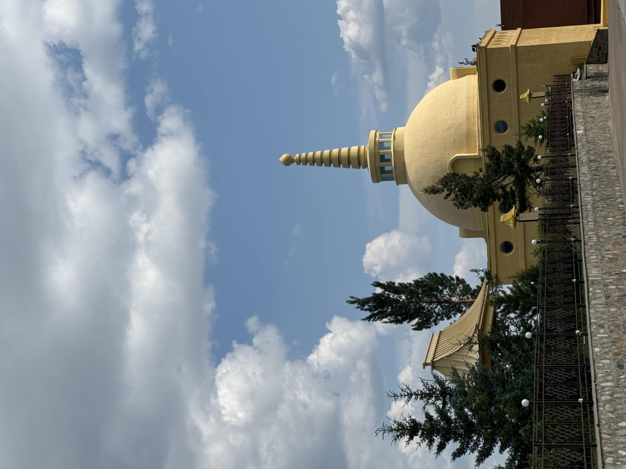 This is a medium-angle, eye-level, outdoor, daylight shot of the Buddhist Temple (Datsan) in Ulan-Ude, Russia. The temple is golden yellow with a large domed roof topped by a spire. A section of the temple building is visible, with several round windows. Several evergreen trees are in front of the temple, and a dark metal fence runs across the foreground. A stone wall is visible below the fence. The sky above is blue with scattered white clouds. A small statue of a figure is visible on the roof of the temple.