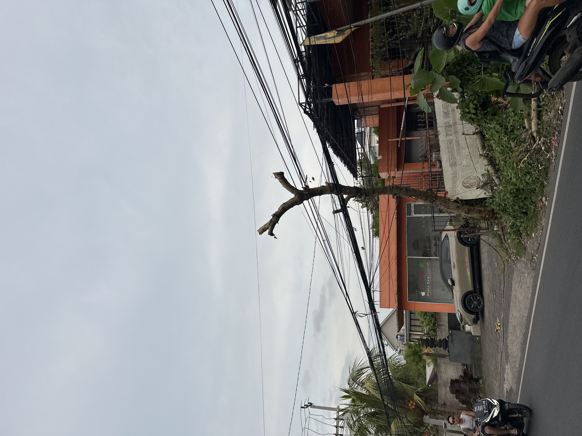 The image, taken in Ungasan, Indonesia, shows a street scene under an overcast sky. A dead tree with bare branches stands near a cluster of electrical wires. An orange building with the sign "Phone Case" is visible, and a light-colored SUV is parked in front of it. A motorcycle with two people wearing helmets is on the right side of the image, while another motorcycle with a single rider wearing sunglasses is on the lower right side of the frame. Lush green vegetation lines the building's edge and the street. A section of the road occupies the lower right corner of the picture. A concrete structure with decorative elements is also observable near the building.
