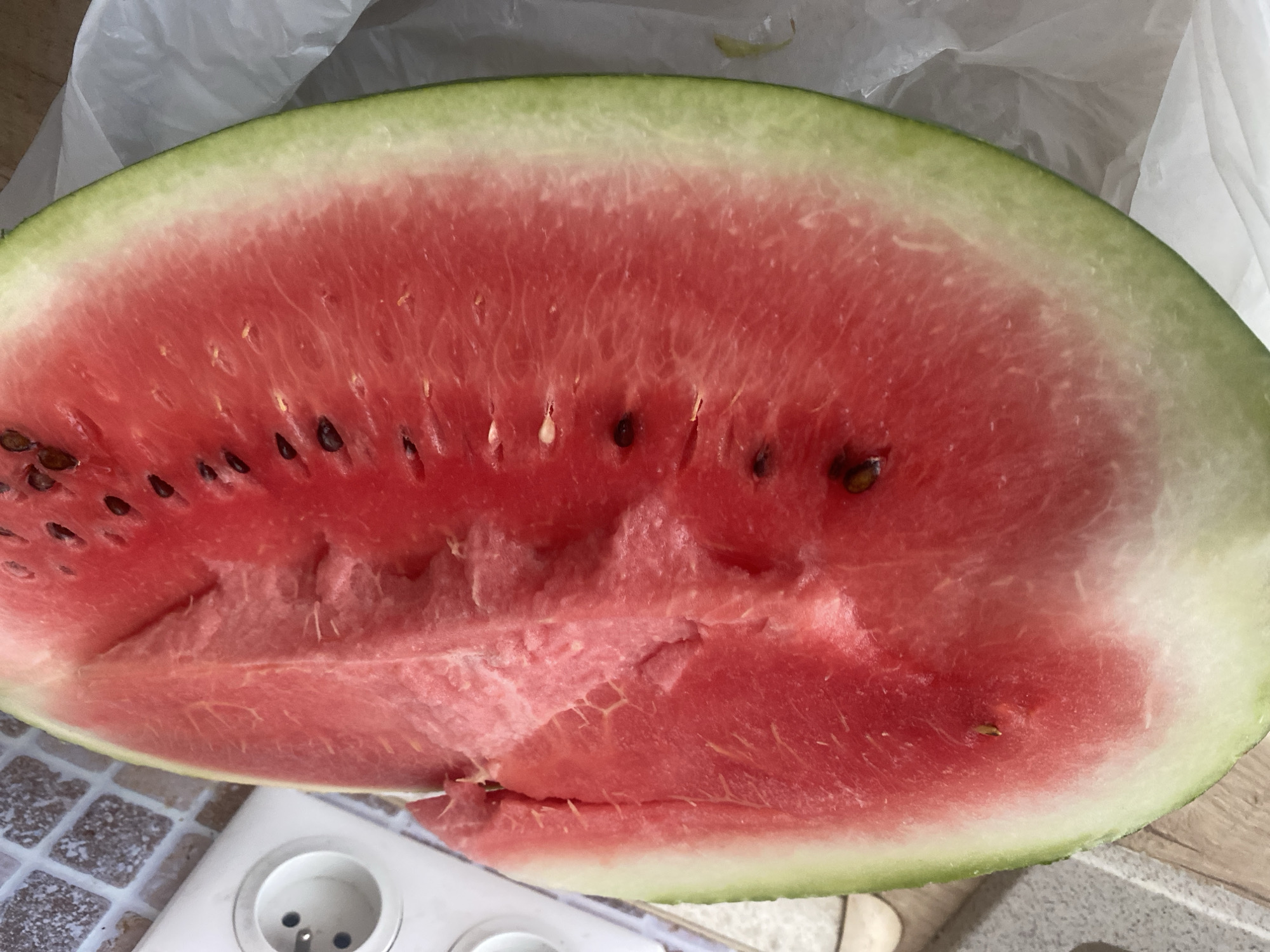 A close-up, overhead shot shows a slice of watermelon placed on a table, partially wrapped in a white plastic bag. The watermelon is vibrant red with black seeds scattered throughout its flesh. The rind is a light green color. To the left of the watermelon, a white electrical outlet is visible on a tiled surface. The scene appears to be an indoor setting, possibly a kitchen. There is no indication of the specific location within Chișinău, Moldova.
