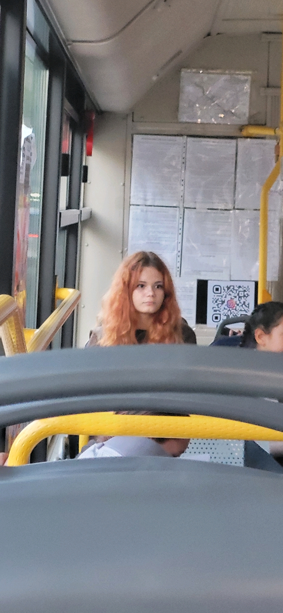 A young woman with long, wavy, reddish-blonde hair sits on a bus. She is looking slightly to her right. She is wearing a dark jacket. Behind her, there are several sheets of paper taped to the wall, and a QR code is visible. Another person's head is partially visible to her right. The interior of the bus is visible, including yellow handrails and gray seats.