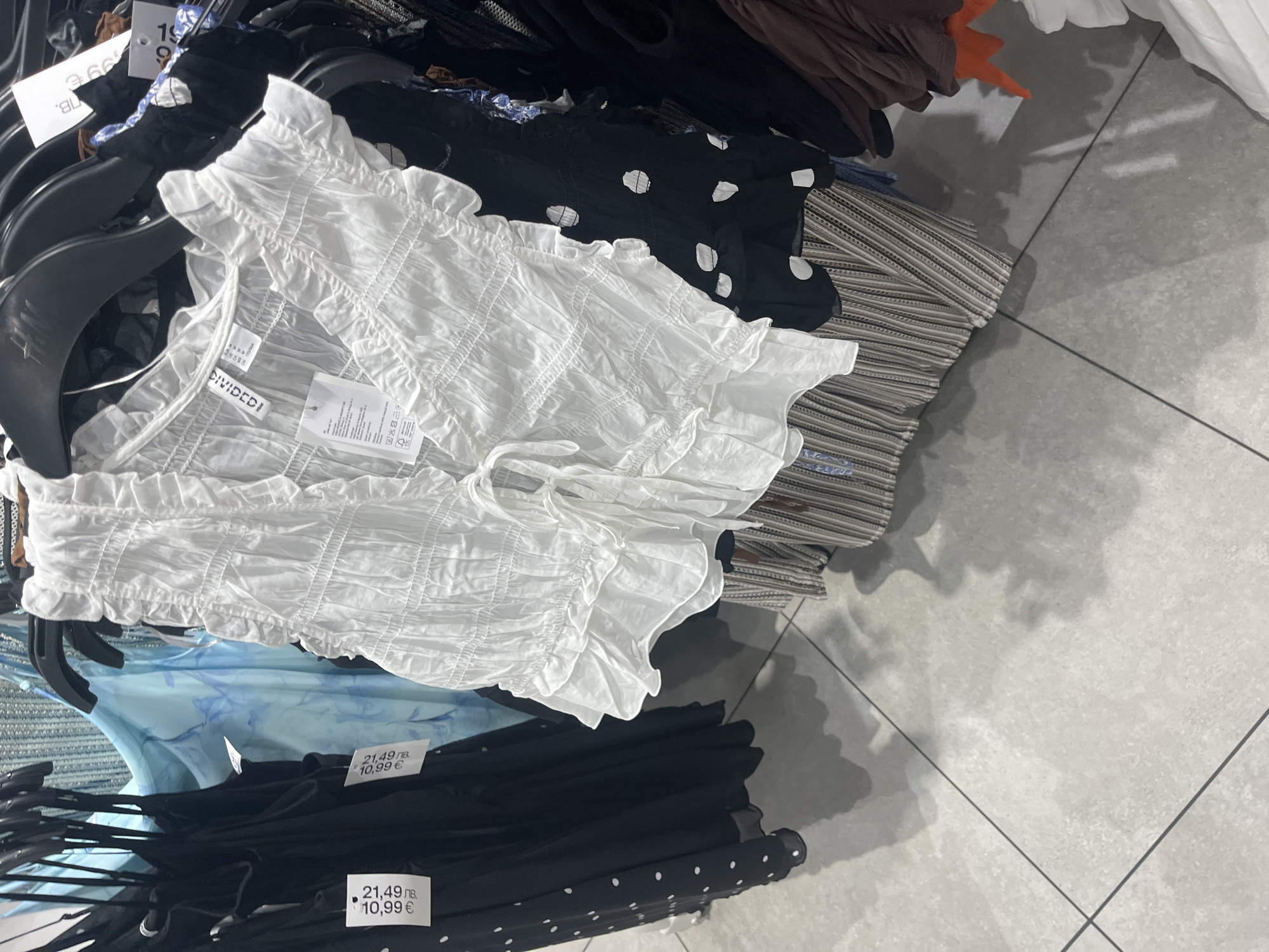 This is an indoor, eye-level shot of clothing on hangers in what appears to be a retail setting. A white, textured, crop top with ruffled edges and ties is prominently displayed on a black hanger. The brand name "DIVIDED" is visible on a tag attached to the garment.
Beneath the white top, there are glimpses of a light blue garment with a possible print, as well as a black piece of clothing with white polka dots. These are also on hangers. Price tags are visible on some of the items, displaying a price of "21.49 лв." and "10,99 €".
Behind the white top and to the right, there's a stack of horizontally striped fabric. Above the white top, there are additional clothes on hangers, with a mix of dark and brown garments.
The floor is tiled in a light gray color, and the overall lighting suggests a retail environment. There is no evidence that this photo was taken in Blagoevgrad, Bulgaria.