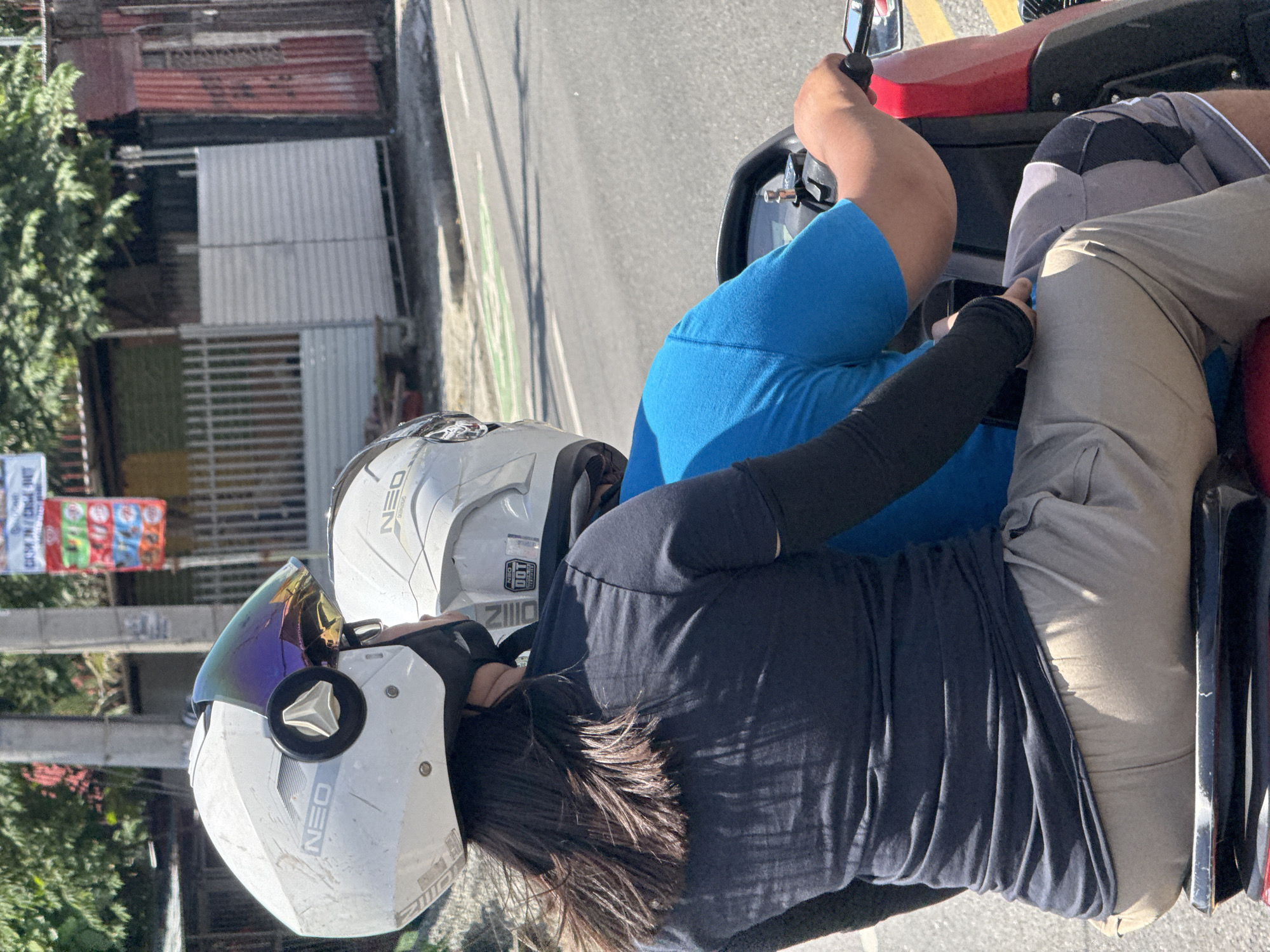 This image, rotated 90 degrees clockwise, depicts two people riding a red and black motorized tricycle, a common mode of transport in the Philippines, on a sunny day in what appears to be an urban or suburban setting in Davao City.
The driver, positioned towards the front, is seen from the back, wearing a bright blue t-shirt and holding the handlebars. Their face is not visible. The passenger, seated directly behind the driver, is dressed in a dark blue or black long-sleeved shirt and light tan or khaki pants. They are wearing a white full-face helmet with a vibrant rainbow-tinted visor and a black face mask covering their lower face. Dark hair is visible spilling from beneath the helmet. Both white helmets feature "NEO" branding, and one also shows "ZITO" and "DOT" marks, indicating safety certification.
The scene includes a concrete road with visible painted lines, including a distinct green line possibly indicating a bike lane, and yellow road markings. In the background, various buildings with corrugated metal roofing and walls are visible, alongside scattered green foliage from trees or bushes. A blurry, colorful banner, likely a political poster given the context, can be seen in the distance on the left. The strong shadows cast by the bright sunlight suggest it is either morning or afternoon. The subjects appear to be in transit, moving along the street.