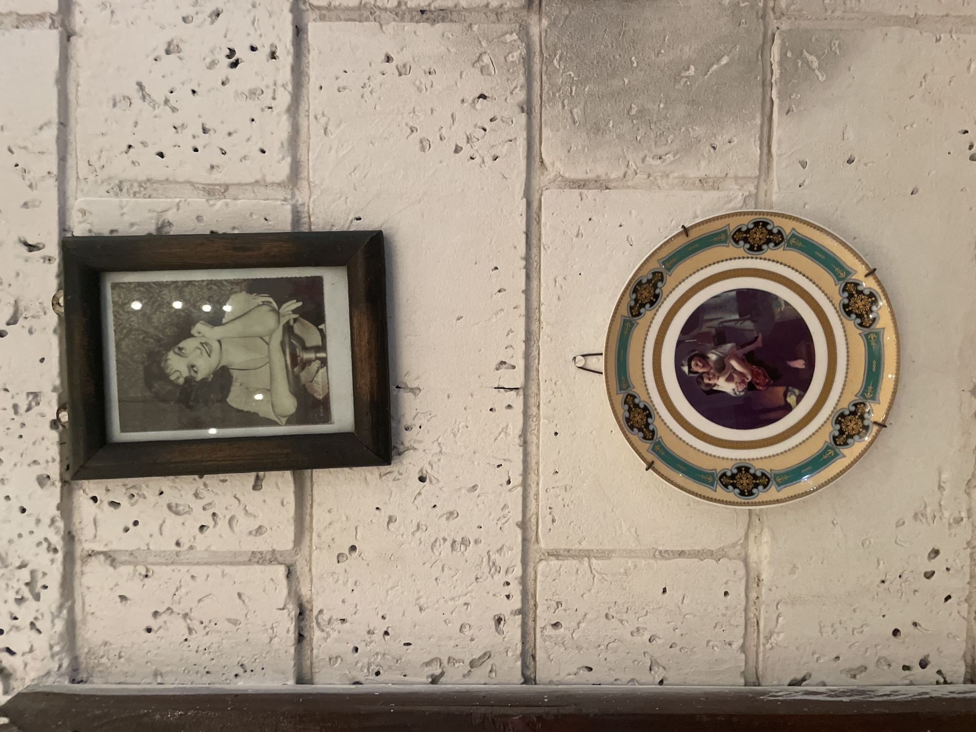 A framed black and white photograph of a woman wearing a dress and resting her chin on her hand hangs on a white brick wall. Below the photograph, a decorative plate with a gold, green, and blue patterned rim hangs on the same wall. The plate features a colorful depiction of two figures in its center. The wall is made of rough-textured, light-colored bricks. A wooden pillar is partially visible on the left side of the image.