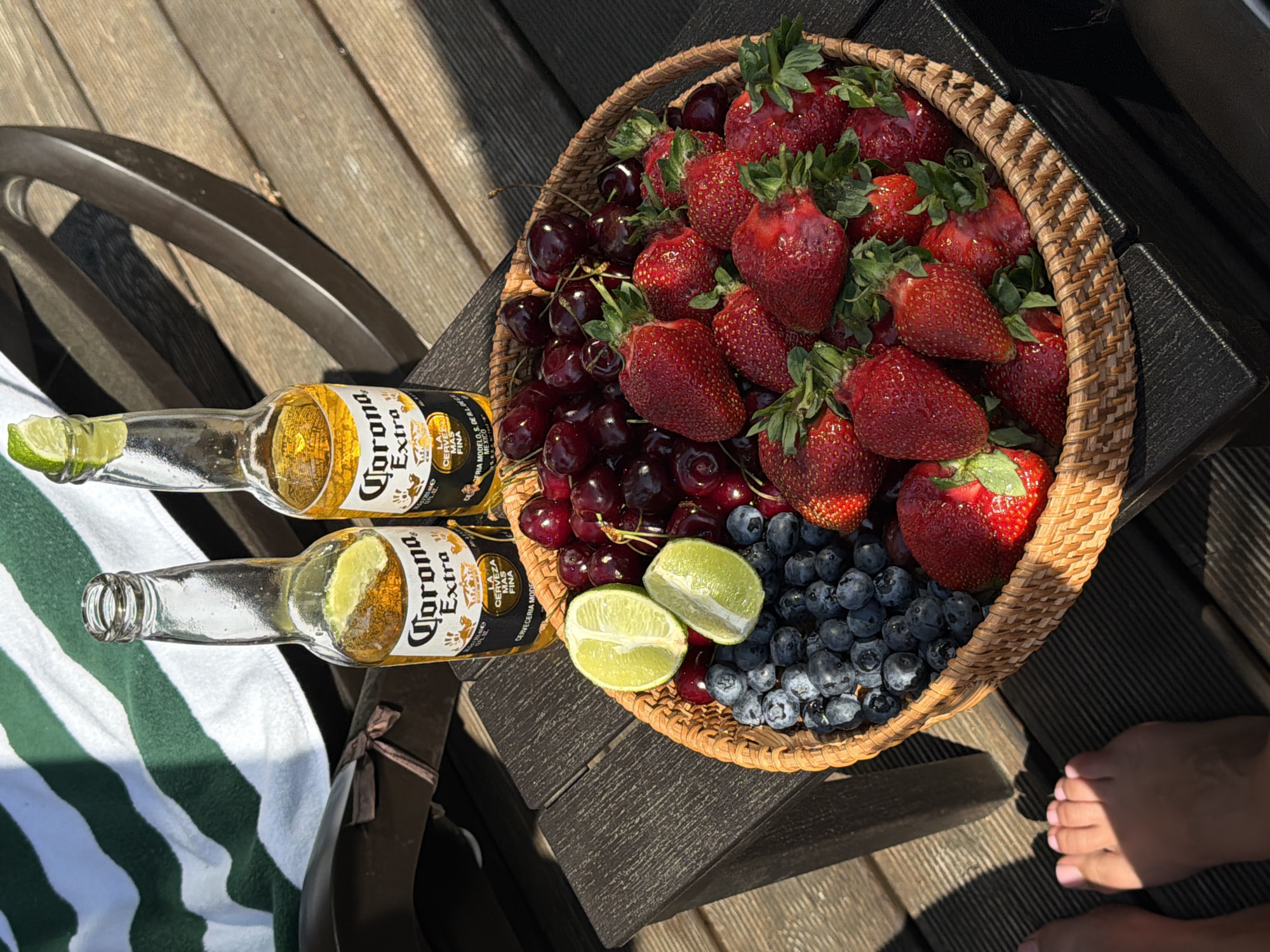 The scene depicts an outdoor setting in Манский, Russia, under direct sunlight, indicated by sharp shadows. A dark, textured outdoor table, likely composite material, holds two open glass bottles of Corona Extra beer, each containing a lime wedge. To the right of the beers, a round, woven basket is filled with fresh fruit: two lime halves, a pile of blueberries, and numerous red cherries and strawberries. In the background, a white towel with green stripes is draped over an unseen object to the left, and a dark, curved furniture frame is partially visible to the right. The entire arrangement rests on a dark wooden deck. In the lower-left foreground, two human feet with light pink painted toenails are visible on the deck, partially in shadow.