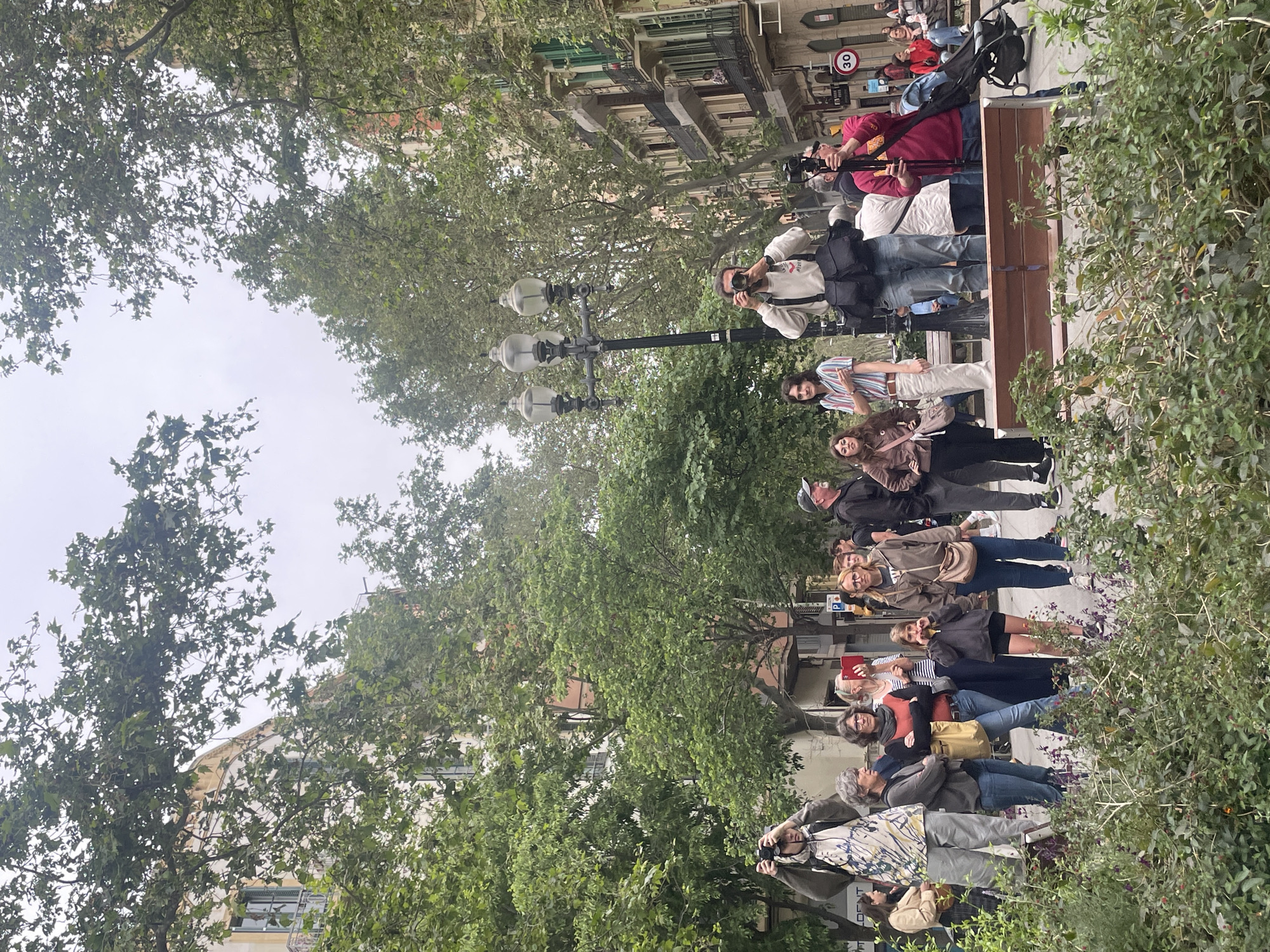 An outdoor, daytime scene captures a diverse group of people gathered along a tree-lined urban path, likely in Barcelona, Spain. The sky is overcast, providing soft, diffused lighting across the scene.
Approximately fifteen to twenty individuals, predominantly adults, are visible, standing along a raised pedestrian area next to a wooden bench. Many are focused on an unseen point to the left of the frame, suggesting they are observing or documenting an event. Several individuals are actively engaged in photography; one person in a red shirt operates a camera on a tripod, and another, wearing a light-colored top, holds a DSLR camera. A third person is seen holding up a smartphone, possibly filming or taking a picture. A baby stroller is visible near the tripod.
The setting is characterized by abundant greenery, with a dense canopy of trees overhead and thick bushes lining the path on the right. In the background, multi-story buildings with balconies and classic architectural details common in European cities are visible.
Notable objects include a tall black lamppost with three lights on the left, a circular "30" speed limit sign, and a blue "P" parking sign in the background. Faint text reading "DENT" is partially visible on a sign in the lower left portion of the image. The general mood of the individuals appears calm and attentive.