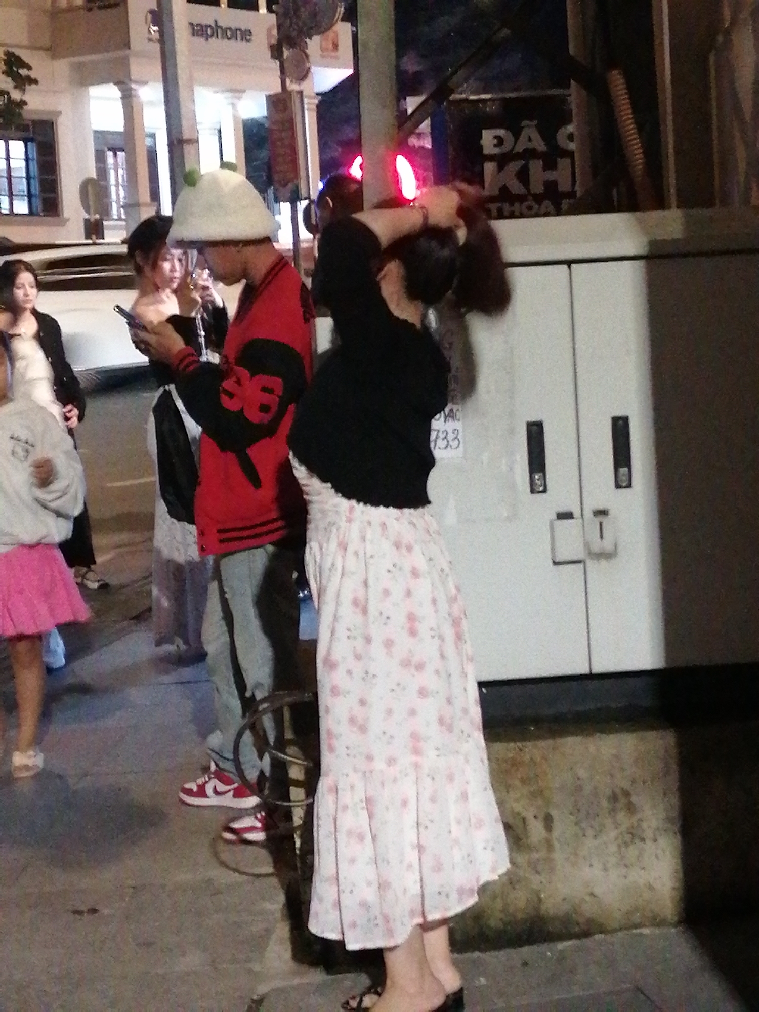 The image depicts a street scene in Da Lat, Vietnam, at dusk. In the foreground, a person wearing a white hat with green pom-poms and a red jacket with black sleeves is looking at their phone. They are standing next to another individual in a black top and a floral skirt. A third person, wearing a gray sweatshirt and a pink skirt, is standing to the left, also looking at a phone. Behind them, a sign for "Nphone" is visible, along with a building with classical architectural elements. The overall environment suggests an urban setting, possibly near a commercial area, given the signage and streetlights.