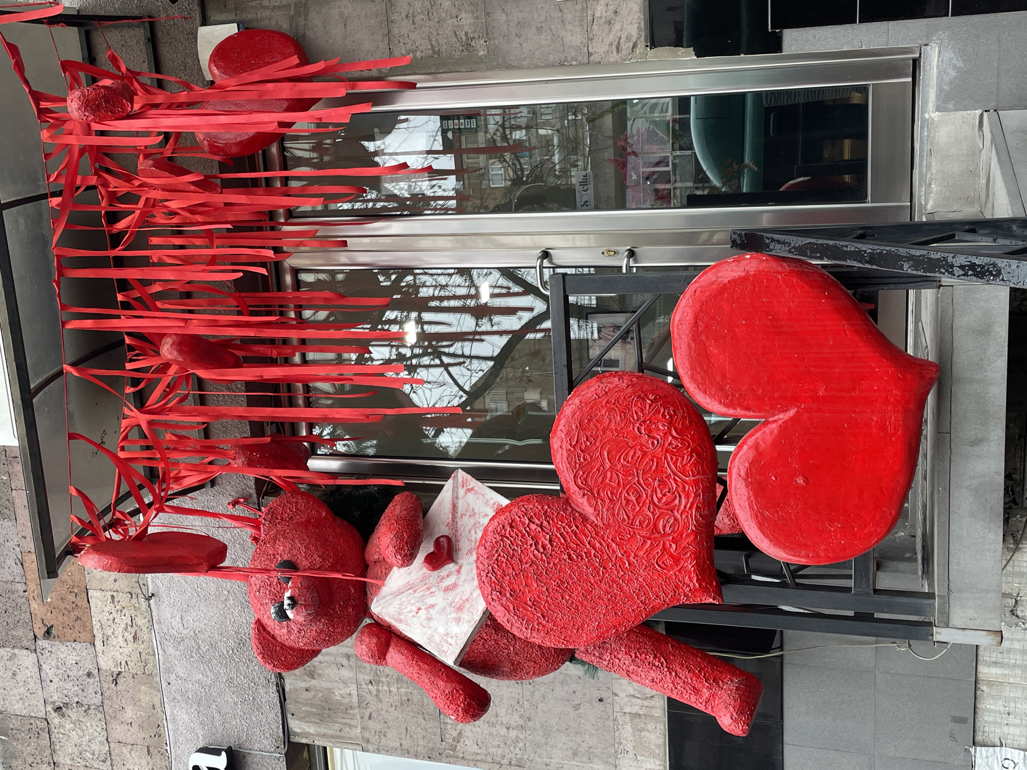 A commercial entrance in Yerevan, Armenia, features a large, red, textured teddy bear statue positioned on a raised concrete platform. The bear holds a white envelope with a small red heart emblem. Below the bear, two large, red, textured heart sculptures rest on the platform. An awning above the entrance is festooned with numerous hanging red ribbons and several smaller red heart ornaments. The entrance consists of double glass doors with metal frames, reflecting bare tree branches and distant buildings. The building's facade displays light-grey textured stone on the left and darker paneling on the right. A black metal railing borders the platform leading to the doors. A faint "Stella" logo is visible on one of the glass door panels. No individuals are present in the scene.