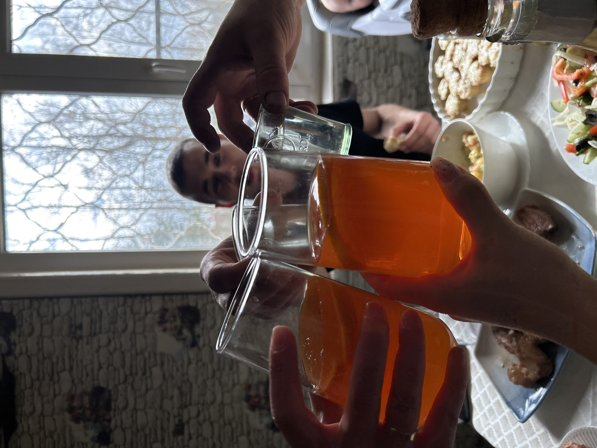 The image captures a moment of celebration, likely a toast, at a dining table inside what appears to be a home. Two people are holding up glasses filled with a bright orange liquid, possibly juice or a cocktail, in a toasting gesture. The hands holding the glasses are prominently featured. The table is set with various dishes, including what looks like salad, potatoes, and meat, suggesting a meal or gathering. In the background, a man's face is visible, partially obscured, indicating he is part of the group. A window provides natural light, revealing a glimpse of the outside. The interior design suggests a home. There is no city specified.