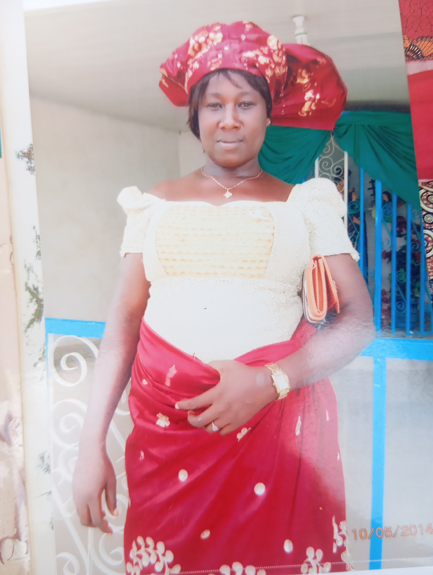 A medium-shot image shows a woman in traditional attire standing outdoors in Karsana 2, Nigeria, with a date stamp "10/05/2014" visible in the lower right corner. She is wearing a red and gold patterned headwrap, a cream-colored off-the-shoulder top with intricate lace detailing, and a vibrant red skirt embellished with gold floral patterns. The woman has a gold necklace with a small pendant and a gold wristwatch on her left wrist. She is holding a small orange clutch bag in her right hand, which is tucked under her left arm. The background features a light-colored wall, a blue railing, and green draped fabric. The overall scene suggests a festive or formal occasion.