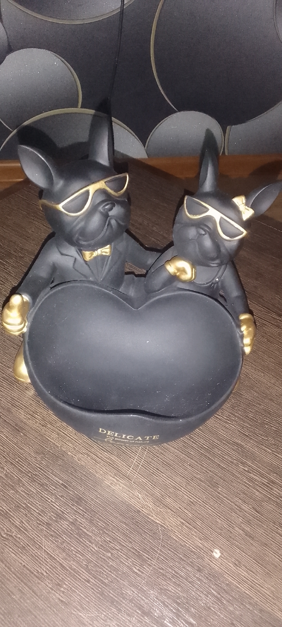 A black, heart-shaped dish is held by two black bulldog figurines wearing gold sunglasses and gold accents. The bulldog on the left is wearing a gold bowtie and a black suit jacket, while the bulldog on the right is wearing a gold ribbon on its head and overalls. Both figurines have their paws on the edge of the dish. The dish has the word "DELICATE" and "The Power of nature" inscribed on it in gold lettering. The background consists of a dark, textured wallpaper with circular patterns. The scene is set on a wooden surface. The location is specified as Topar, Kazakhstan.