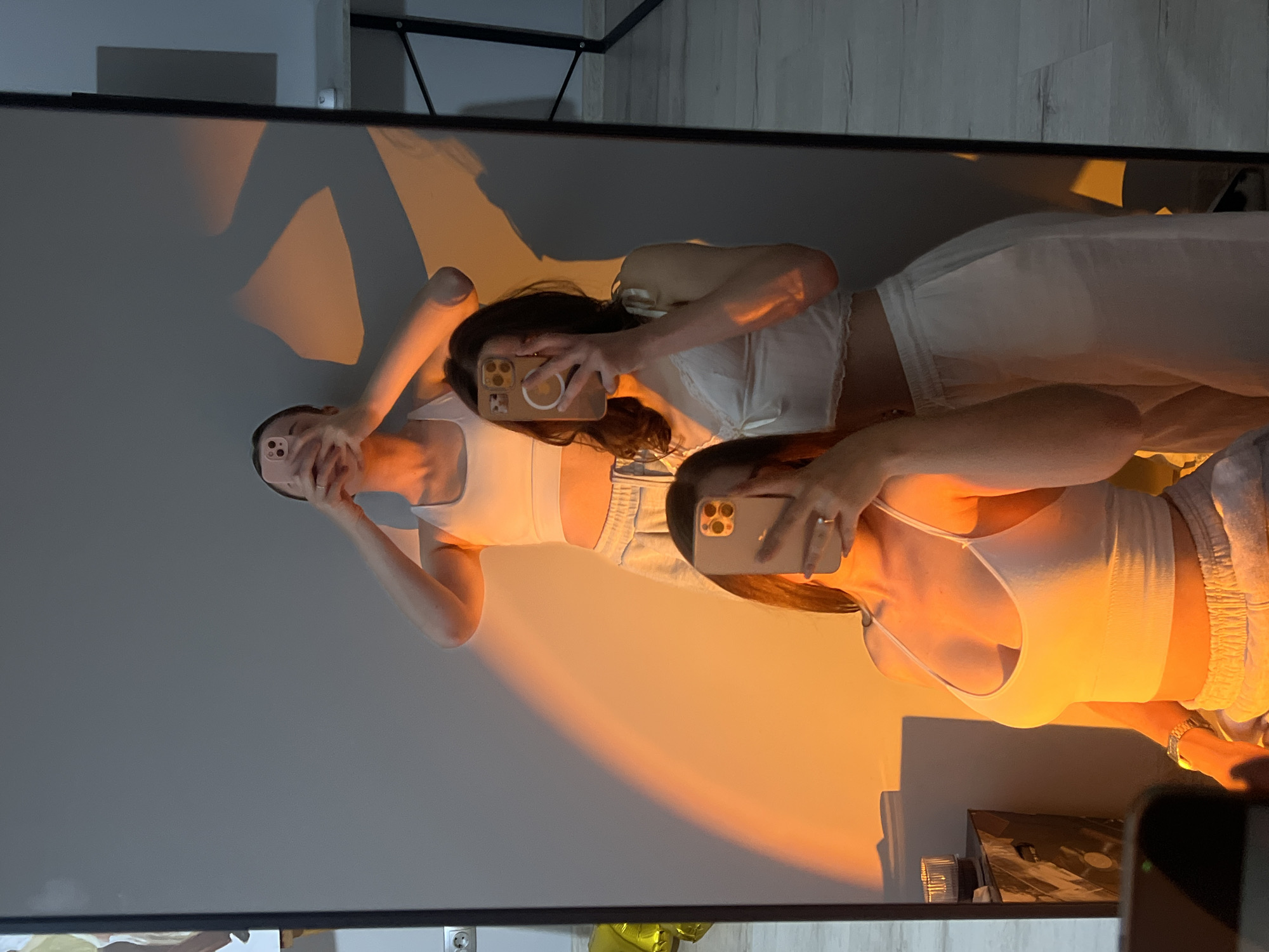 Three young women are taking a mirror selfie in an indoor setting, possibly an apartment. They are all dressed in casual, light-colored loungewear. The woman in the foreground is wearing a white tank top and light-colored pants, and is holding her phone to take the picture. The woman behind her is wearing a similar white top with delicate lace trim and matching pants, also holding a phone. The woman in the back, further up, is wearing a white sports bra and pants and is holding her phone at a higher angle. A warm orange light casts a dramatic glow across the wall behind them, creating interesting shadows. In the reflection of the mirror, part of a desk and a wall with electrical outlets are visible. The floor appears to be made of light-colored wood planks. There is a yellow balloon visible on the left side of the image, partially in the reflection. The overall impression is a candid, casual moment captured in what seems to be a well-lit room. The specific location in Ternopil, Ukraine cannot be determined from the image.