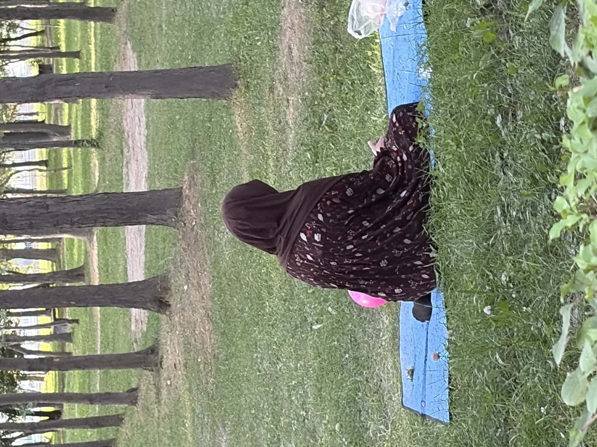 A person wearing a dark brown hijab and a dark floral print dress sits on a blue mat in a grassy area with trees in the background. The person is facing away from the camera, with their left arm resting on their lap. A bright pink balloon is partially visible next to the person. A white plastic bag is visible to the right of the mat. The environment appears to be a park or wooded area with a dirt path visible in the distance.