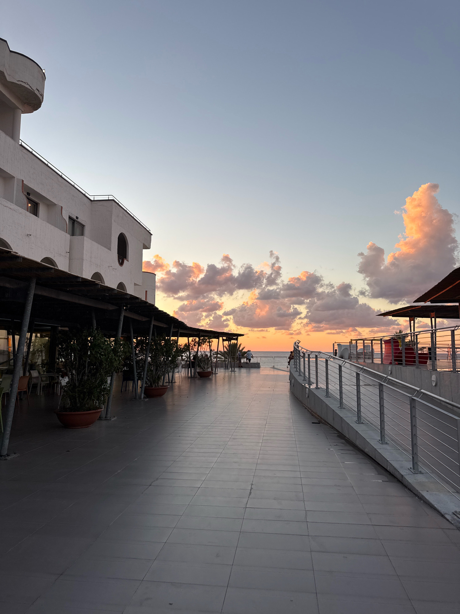 This image captures a sunset scene on a wide, tiled promenade in Isola delle Femmine, Italy. On the left, a multi-story white building with arched windows and a curved balcony overlooks the promenade. A covered walkway with metal supports extends along the building, sheltering tables and chairs. Large potted plants with dark green foliage are arranged along the edge of the walkway.
The promenade itself is paved with large, square grey tiles. A metal railing with horizontal bars runs along the right side of the promenade, leading down a ramp. Beyond the railing, there appear to be outdoor seating areas with tables and chairs, some with red elements. In the distance, the sea is visible, meeting the horizon under a sky filled with soft orange and pink clouds, illuminated by the setting sun. Two figures can be seen in the distance on the left side of the promenade near the water's edge. The overall atmosphere is one of a tranquil evening by the sea.