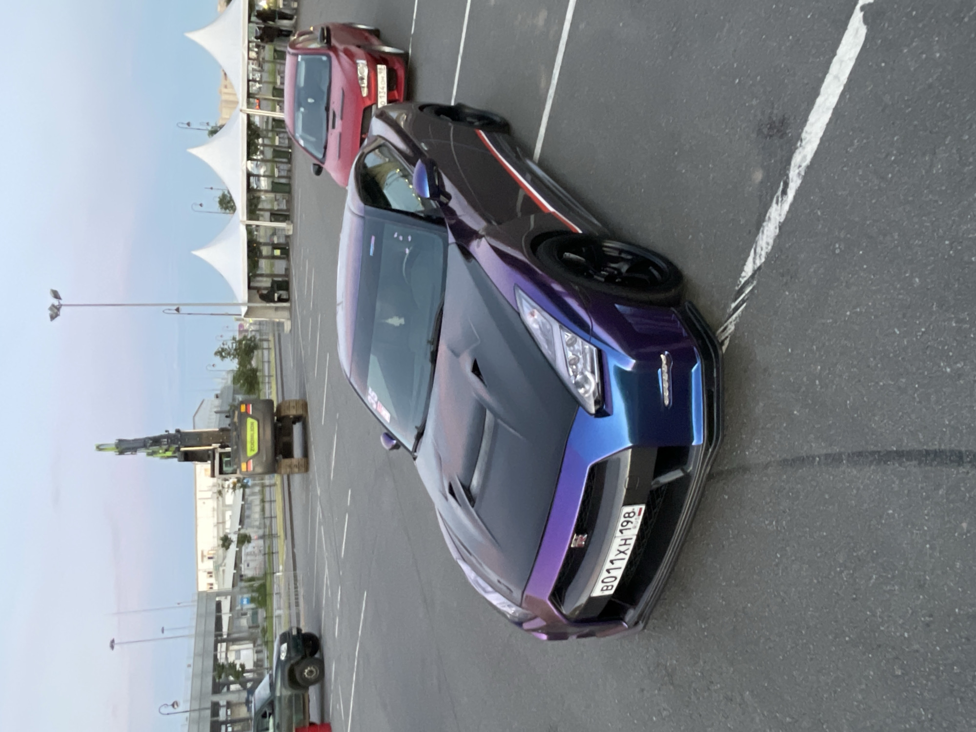 The image captures an outdoor scene at what appears to be an event or parking lot, with a focus on a customized purple and blue Nissan GT-R parked in the foreground. The car has a distinctive iridescent paint job and racing stripes on its side. A red Mitsubishi Lancer is parked behind it, and further in the background, a dark green SUV is visible. An excavator is positioned in the mid-ground to the left, and several white tent-like structures are set up to the right, suggesting an event or venue. The ground is paved asphalt with visible parking lot lines. The sky is a light blue with some clouds, indicating daytime. The overall environment suggests an urban or semi-urban setting.