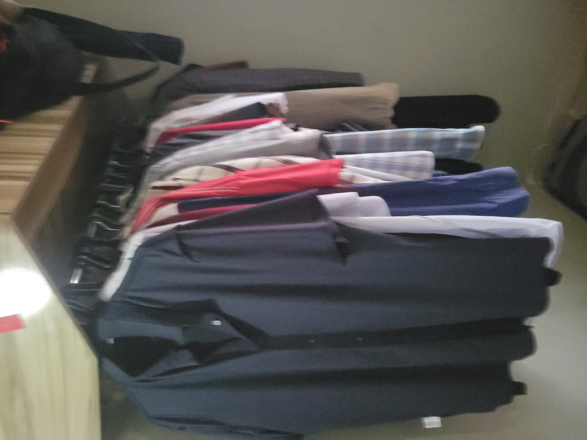 A close-up, dimly lit, slightly blurry shot shows a collection of clothing hanging on a rack. The most prominent item is a dark blue or black short-sleeved collared shirt in the foreground, hanging on a black hanger. Behind it, other garments are visible, including red, white, and plaid shirts, as well as some neutral-colored items. The rack itself appears to be a simple metal rod. Above the clothes, a wooden shelf or plank is visible, with a bright light source illuminating the area. The background is a plain, light-colored wall. No people or specific location details are discernible.