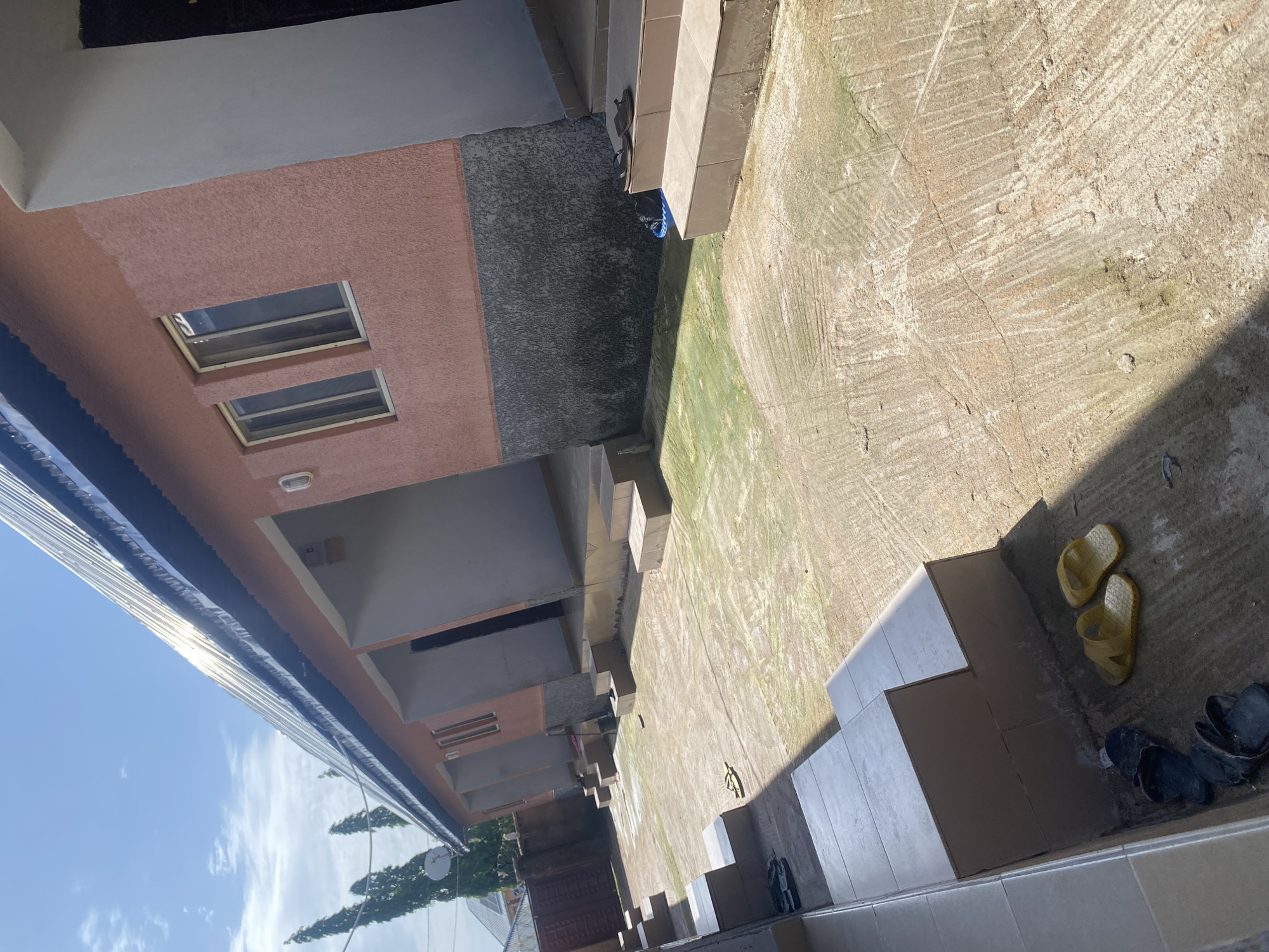 The image depicts an outdoor courtyard area with a building. The building has a peach-colored textured facade with two rectangular windows. Below this, there's a section with a dark, speckled texture and a corrugated metal awning extending over the courtyard. Adjacent to the building, a tiled staircase leads down into the courtyard.
The courtyard floor is a rough, textured concrete surface, showing signs of wear and possibly moss growth in some areas. There are several pairs of flip-flops scattered on the ground near the staircase and a separate set of dark sandals. The overall scene suggests a residential or possibly a commercial building in a tropical or sub-tropical environment.
The lighting indicates it is daytime, with strong shadows cast by the building and other structures, suggesting the sun is at a moderate angle. The sky is visible in the upper left portion of the image, showing blue sky with some wispy clouds, and what appears to be the top of trees and a satellite dish. The perspective is tilted, giving a dynamic feel to the composition.
There is no visible text in the image. The presence of casual footwear suggests a relaxed, everyday setting. The location context of Jalingo, Nigeria, aligns with the visual cues of the climate and architectural style.