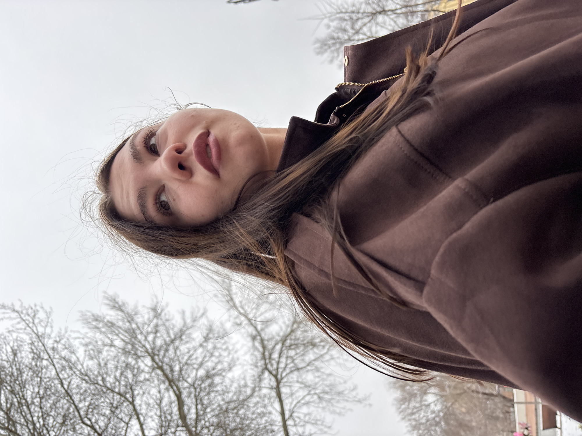 The media file presents a low-angle portrait of a young woman, rotated 90 degrees clockwise. The woman is looking downwards towards the camera with a neutral or slightly thoughtful expression. She has long, dark brown hair, dark eyes, and pronounced eyebrows, and is wearing dark, possibly plum-colored lipstick. She is dressed in a dark brown jacket or coat featuring a visible zipper and a structured collar.
The setting is outdoors on an overcast day, with a bright, diffused light illuminating the scene. The background consists predominantly of a pale, cloudy sky, against which the stark silhouettes of numerous bare deciduous trees are visible, indicating late autumn or winter. A very blurry and indistinct hint of a building or structure and faint pink details can be seen in the far background on the right side. The overall atmosphere is muted due to the cloudy weather. No visible text is present in the image. The location is Perm, Russia.