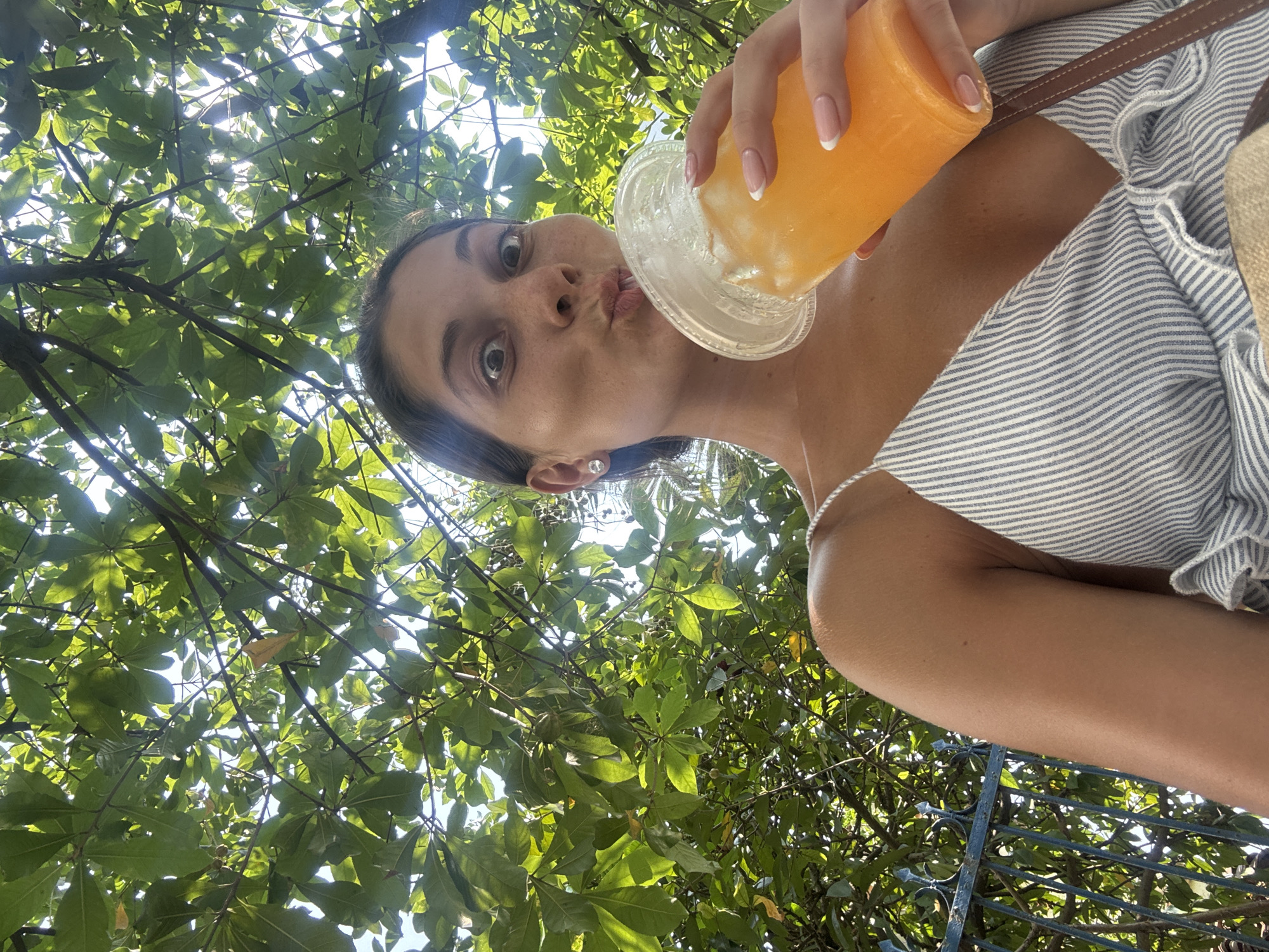 A woman poses for a low-angle selfie with a clear plastic cup filled with an orange beverage and ice. She is wearing a light-colored, striped top with thin straps. The background consists of lush green foliage and glimpses of the sky. Based on the vegetation and architectural styles often observed in the area, it's possible this scene could be located in Đà Nẵng, Vietnam.