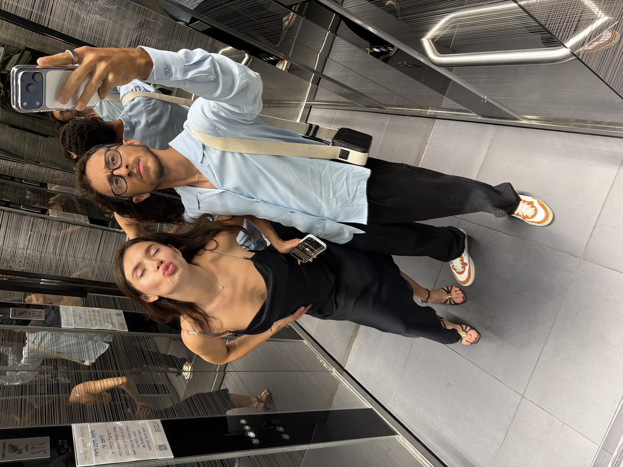 In an elevator with mirrored walls and brushed metal accents, a young couple takes a selfie. The woman, on the left, wears a black slip dress and strappy heels, puckering her lips for the camera. The man, on the right, sports a light blue button-down shirt, black pants, and sneakers. He holds a smartphone in his left hand, capturing the moment. A black crossbody bag hangs from his shoulder. The elevator floor is tiled in a neutral gray. Reflections of other elevator doors and signage are visible in the mirrored surfaces.