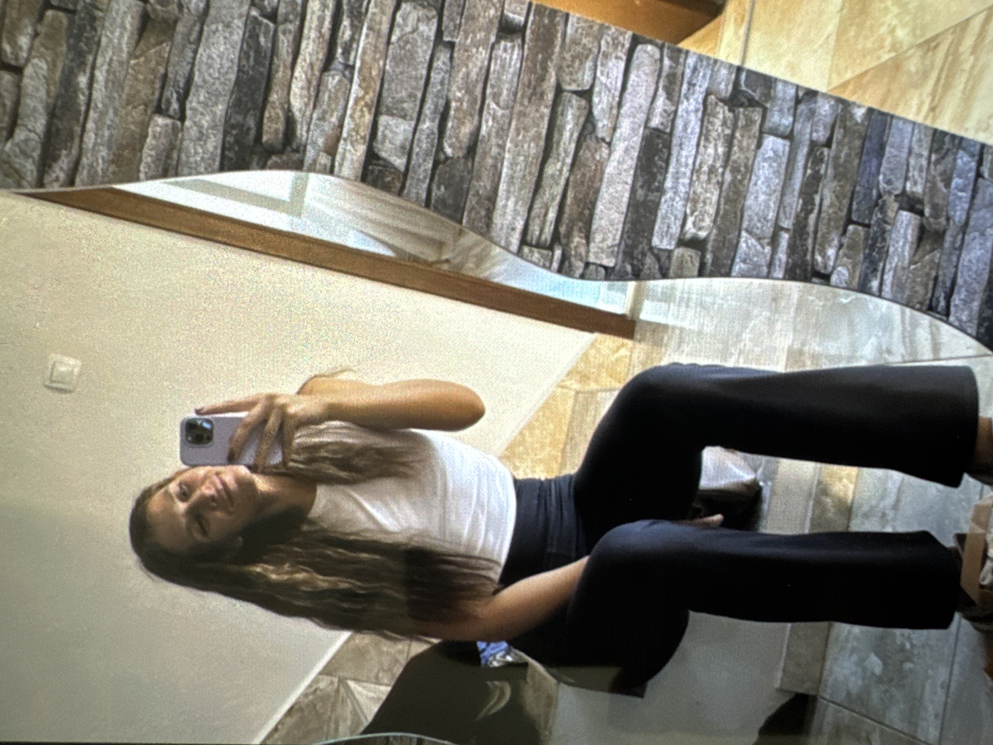 A female individual is depicted in an indoor setting, identified as being located in Vratsa, Bulgaria. She is seated, facing a mirror, and holding a light purple smartphone in her right hand, apparently capturing a mirror selfie. Her head is slightly tilted, and she exhibits a subtle smile. She possesses long, wavy brown hair that extends over her shoulders. Her attire consists of a white sleeveless crop top and dark, high-waisted, wide-leg trousers. Her left arm rests on her left leg. The immediate environment includes a tiled floor and steps beneath and around her. The reflected background in the mirror reveals a plain, light-colored wall to the left, and a decorative wall section on the right, clad with horizontally arranged stones of various shades of grey, brown, and off-white. Vertical wooden frame elements are visible within the reflected space.