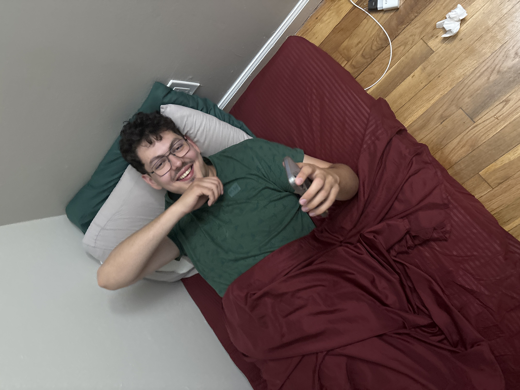 A young man, likely in his late teens or early twenties, is pictured lying on a bed in an indoor setting within Franklin Township, United States of America. He has short, curly dark hair and wears glasses along with a dark green patterned t-shirt. He is smiling broadly, looking directly at the camera, conveying a cheerful and relaxed demeanor.
He is propped up against a light grey wall, resting on a dark green pillow and a light grey/white pillow. His lower body is covered by a maroon or dark red sheet or comforter. In his right hand, he holds a grey smartphone.
The floor adjacent to the bed is made of light-colored wood planks. Visible on the wooden floor is a white charging cable connected to a white wall adapter, which has partially legible text resembling "XIAOMI." A small, crumpled piece of white paper or tissue is also on the floor. A white electrical wall outlet is present on the wall behind the bed. The scene suggests a casual moment of rest or leisure in a bedroom environment.