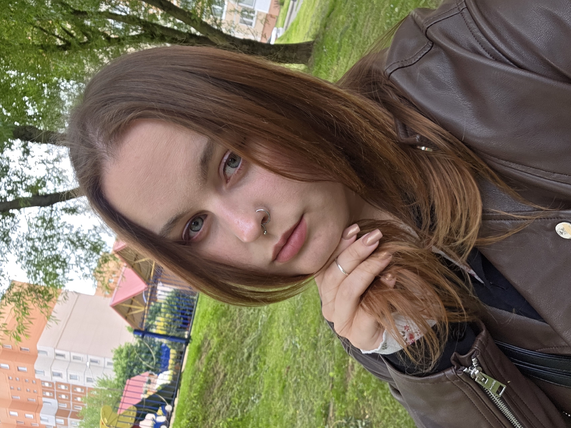 A portrait of a woman is presented outdoors. The woman has fair skin, light eyes, and brown hair. She is wearing a brown leather jacket and has a nose ring on each side of her nose. Her fingernails are painted a light tan color. She is holding a strand of her hair with her right hand, which also has a silver ring on one of the fingers.
The background features a grassy area and a building that appears to be a residential complex with an orange facade. Additionally, there's a playground structure with a red roof visible in the background. The location appears to be in a city park or a recreational area adjacent to residential buildings.