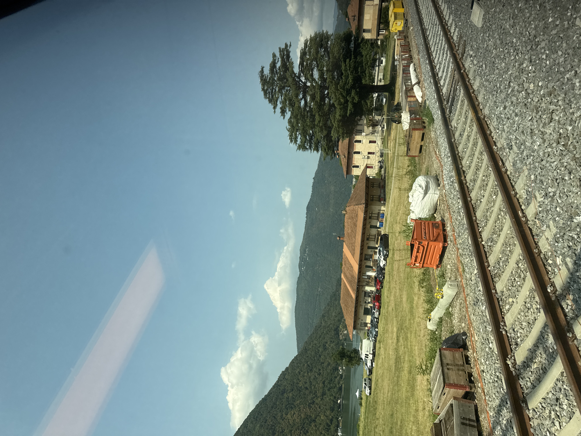 The image depicts a scenic view of Val Mara, Switzerland, on a clear day. The foreground is dominated by a railway track, with ballast and concrete sleepers visible. To the left of the tracks, there are several wooden crates, some empty and others appearing to contain materials, alongside a large white sack. An orange industrial skip is also present.
In the mid-ground, a grassy field stretches towards a low-rise building with a terracotta-tiled roof, likely a train station or a similar facility. A line of parked cars, including a white van and several sedans, is situated in front of the building. Behind the building, a large, mature tree with a broad canopy stands prominently. Further in the background, more buildings and residential structures are visible, nestled against a backdrop of lush green hills and mountains.
The far background features a body of water, possibly a lake or a bay, with a distant shoreline and more forested hills. The sky is a clear blue, with a few scattered white clouds, one of which is particularly large and puffy. A subtle rectangular reflection is visible in the upper part of the image, suggesting it was taken through a window.