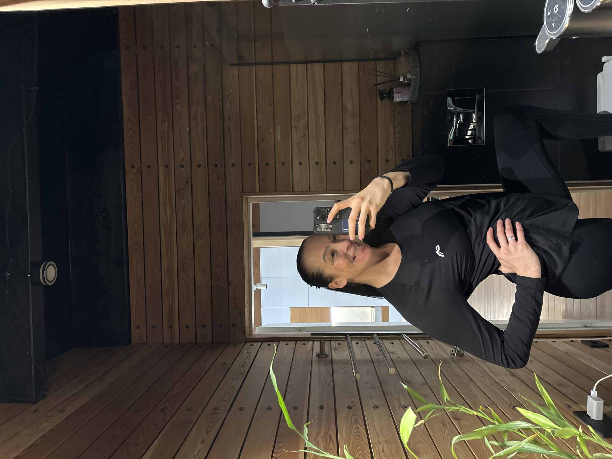 This image, located in Chaikovsky, Russia, depicts a female subject capturing a mirror selfie within an indoor environment. The woman, with dark hair tied back, smiles at her reflection. She is dressed in a black long-sleeved top featuring a white logo on the left chest and black leggings. Her right hand is placed on her hip, while her left hand holds a dark smartphone, pointed towards the mirror. Her left knee is bent, and her left foot is raised.
The immediate background of the room consists of walls covered in horizontal light-brown wooden paneling, secured with visible dark screws. The ceiling above is black and features a single circular recessed light fixture. In the lower left foreground, a green plant with long leaves partially obscures a gray charging adapter plugged into a wall outlet, from which a white cable extends. A three-bar chrome towel rack is mounted horizontally on the wooden wall to the left of the woman.
The mirror's reflection reveals additional elements of the room. To the left of the woman, a light-framed doorway is visible, opening into another room characterized by white walls and a ceiling-mounted light fixture. To the right of the woman's reflection, a dark surface supports a small woven basket containing a dark spray bottle and several diffuser sticks. Below this arrangement, a rectangular chrome flush plate for a toilet is mounted on a dark wall, with a portion of a white toilet bowl discernible beneath it.