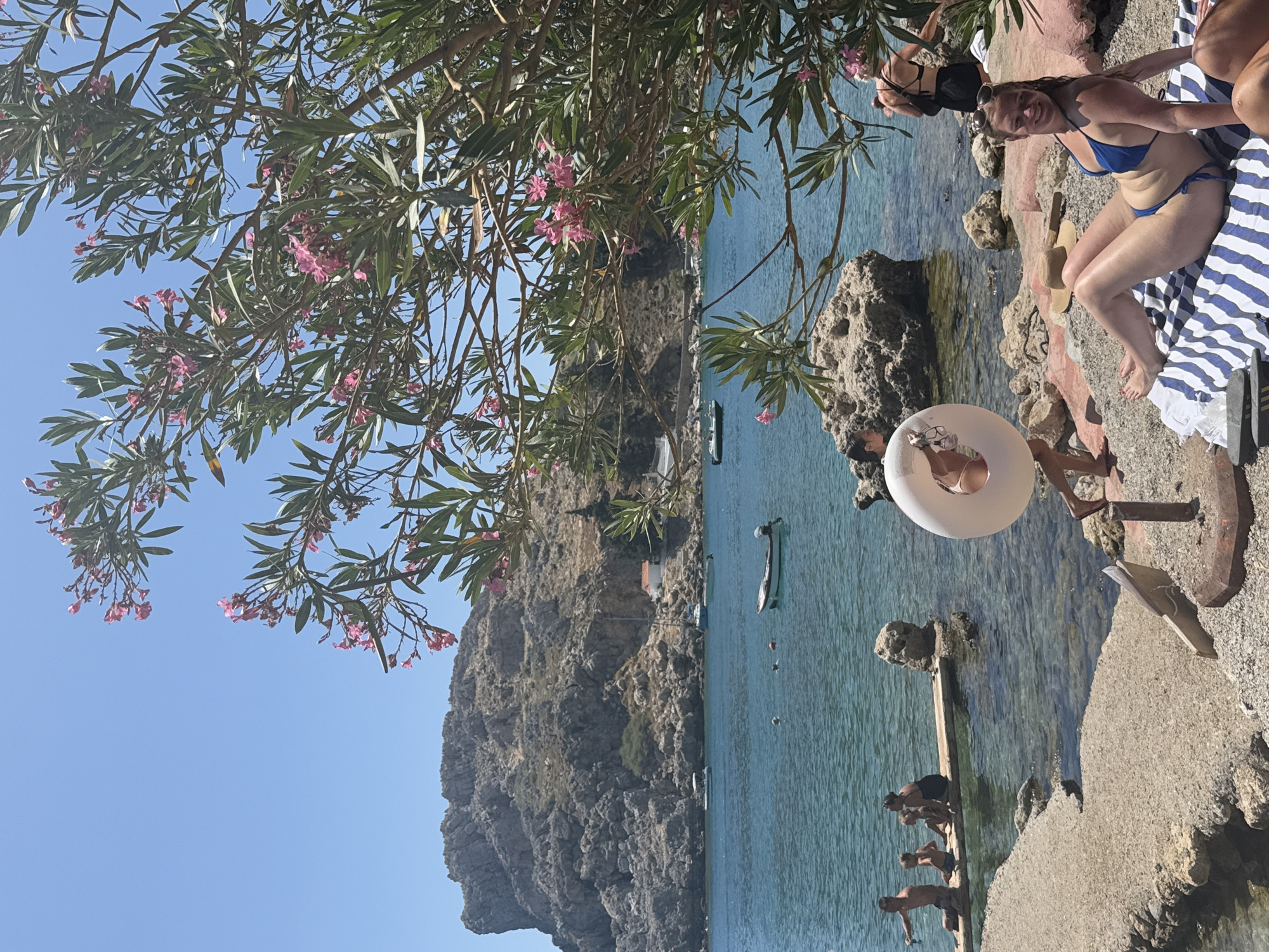 A wide shot captures a picturesque bay in Lindos, Greece, under a clear blue sky. In the foreground, a woman in a blue bikini sunbathes on a striped towel on a pebble beach. To her left, a white inflatable ring is held by a person partially visible, while another person in a black swimsuit stands further back. To the left, a wooden platform extends into the turquoise water, with a family of four, including two children, sitting on it. The water is calm, with a small white boat moored in the middle ground and other boats visible in the distance. A rocky, tree-covered hillside with a small white building and an orange roof dominates the background. Blooming oleander branches with pink flowers frame the scene from the right and upper part of the image.