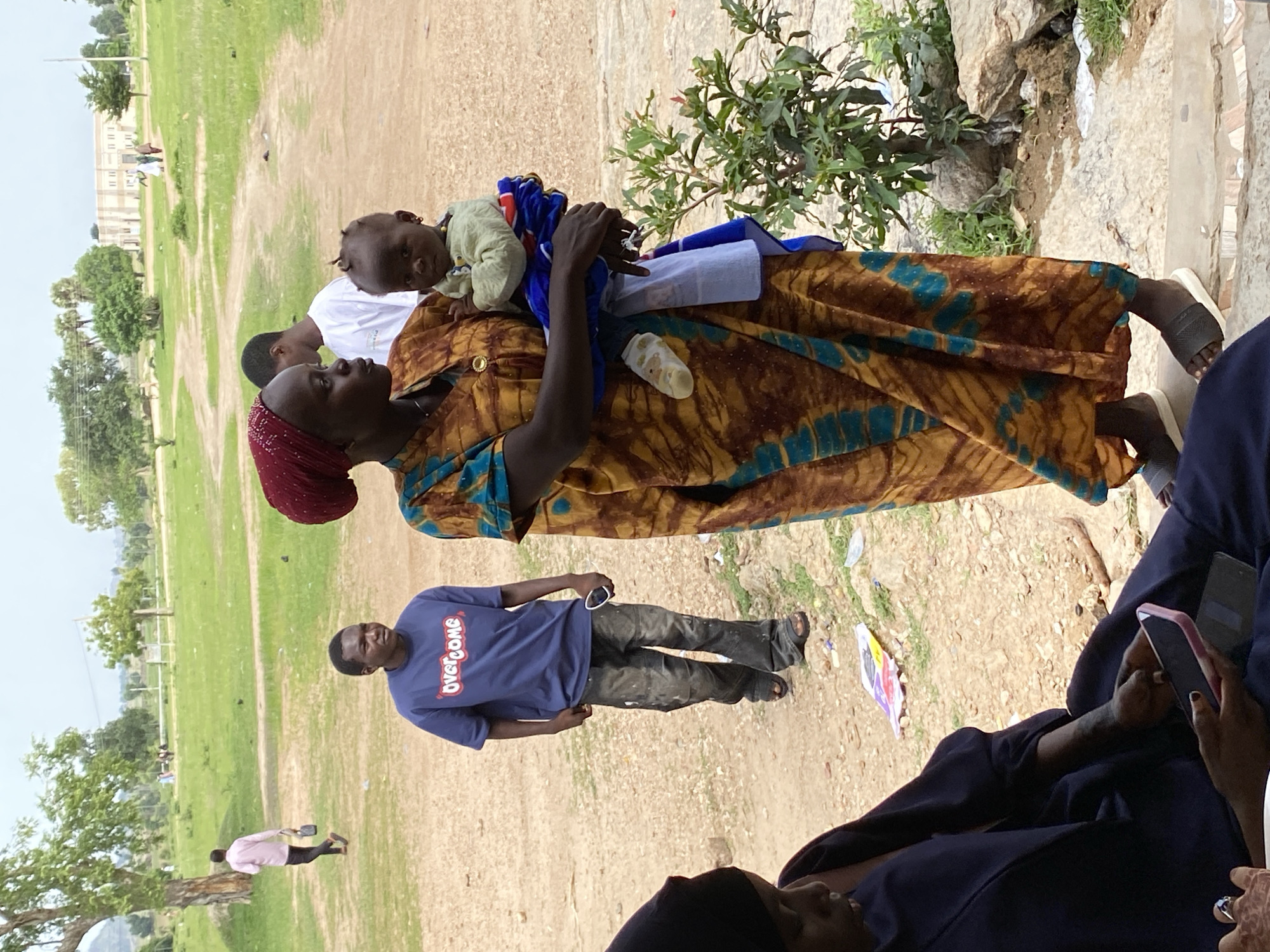In Yali, Nigeria, a woman in a colorful, patterned dress and red headscarf holds a baby wearing a light green outfit. The baby is facing forward. Behind them, a young man in a blue t-shirt with "OVERCOME" printed on it walks away from the camera, holding a white object. Another individual, partially visible on the left, wears a dark blue garment and looks down at a pink-cased phone. The environment is an outdoor, dusty area with scattered green vegetation and rocks. In the background, several people are seen in the distance near a building.