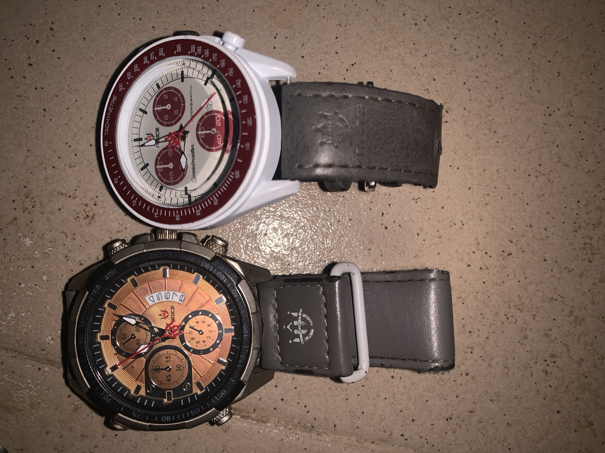 Two wristwatches are displayed side-by-side on a textured surface that appears to be a tiled floor. The watch on the left has a metallic casing and a copper-toned dial with three sub-dials. The watch on the right has a white casing with a red bezel and a white dial featuring red accents and two sub-dials. Both watches have grey leather straps. The left strap features a light-colored emblem. The scene is illuminated by artificial light, casting subtle shadows.