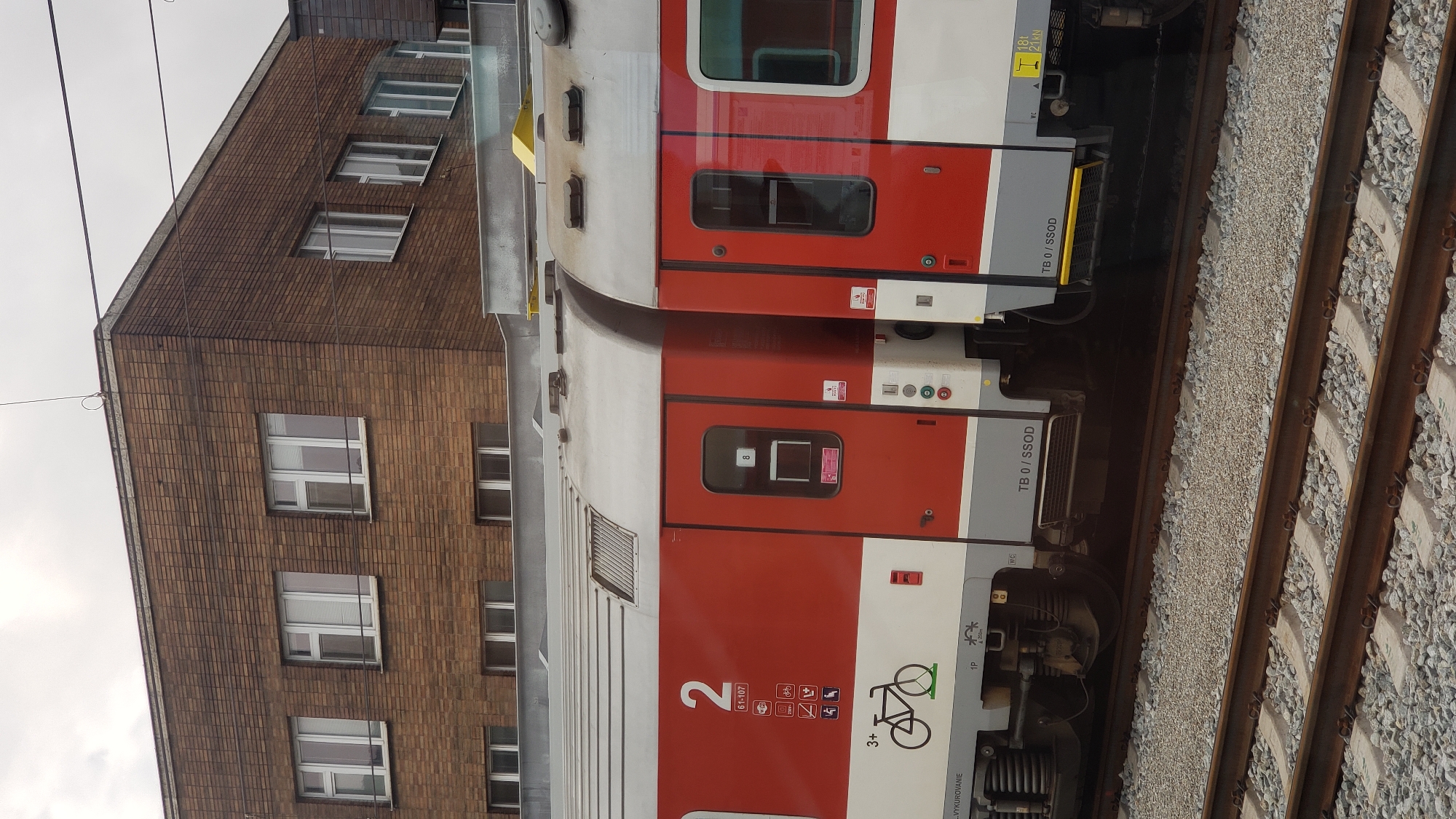 The image shows the side of a red and white train car, with the number "2" prominently displayed. Various symbols, including a bicycle, are visible on the side of the train. The train is on a railway track, with gravel and sleepers visible. In the background, a brick building with multiple windows is seen. The scene appears to be an outdoor railway station. The overall impression is that of a typical urban or suburban railway environment.