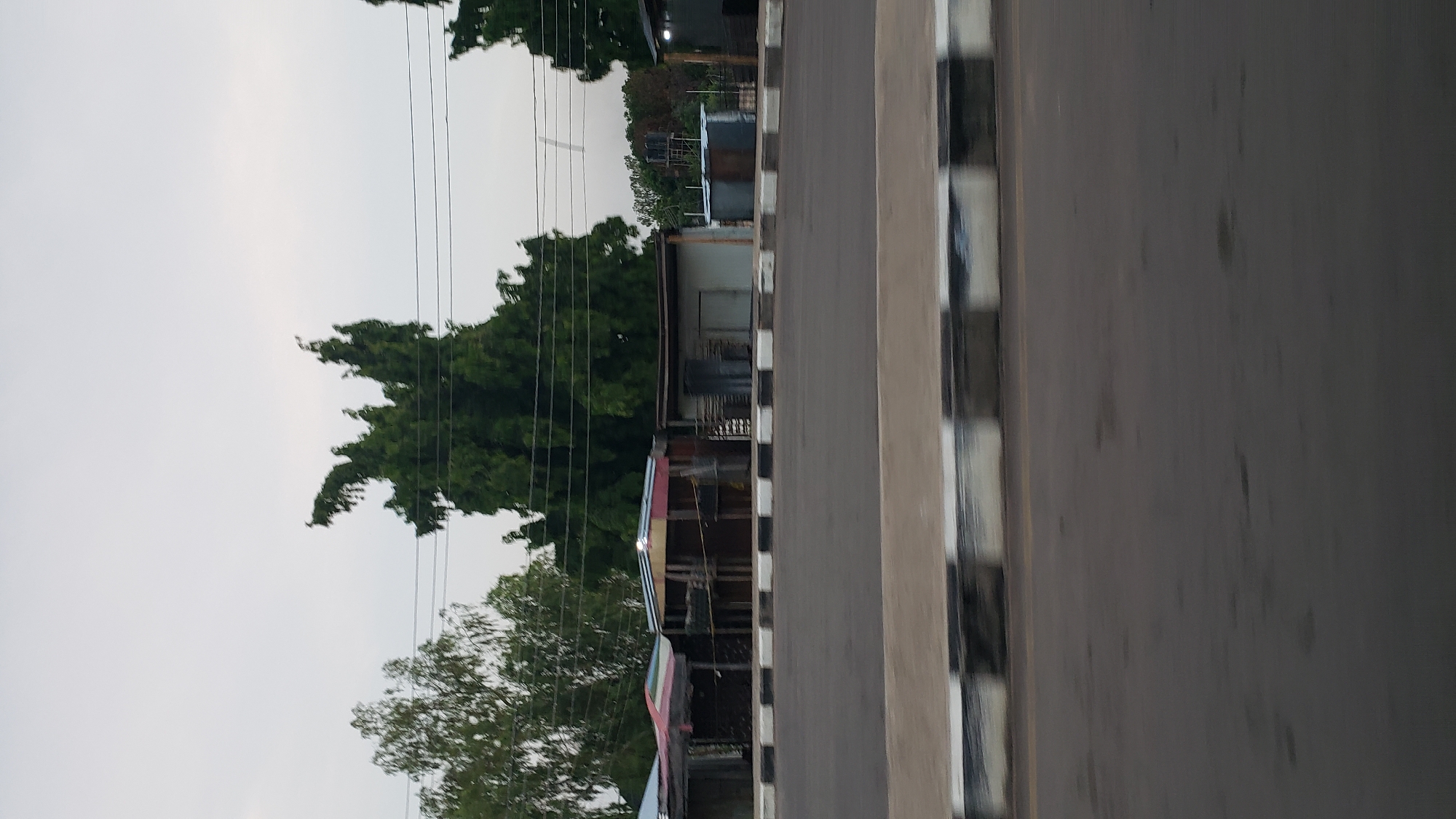 In Jalingo, Nigeria, a low-angle shot captures a row of small, makeshift shops lining a paved road. The shops, constructed from corrugated metal and wood, exhibit various levels of disrepair and unfinished appearances. Some have open fronts, revealing interior structures, while others are closed with doorways. One shop features a striped awning. The shops are situated behind a curb with alternating black and white rectangular blocks, separating them from the road. Trees with dense green foliage are visible behind the structures, and utility wires stretch across the sky. The sky is overcast and a pale gray. The road in the foreground is dark and appears wet in places, with subtle patterns suggesting reflections or puddles. The overall scene depicts a commercial area with basic infrastructure.