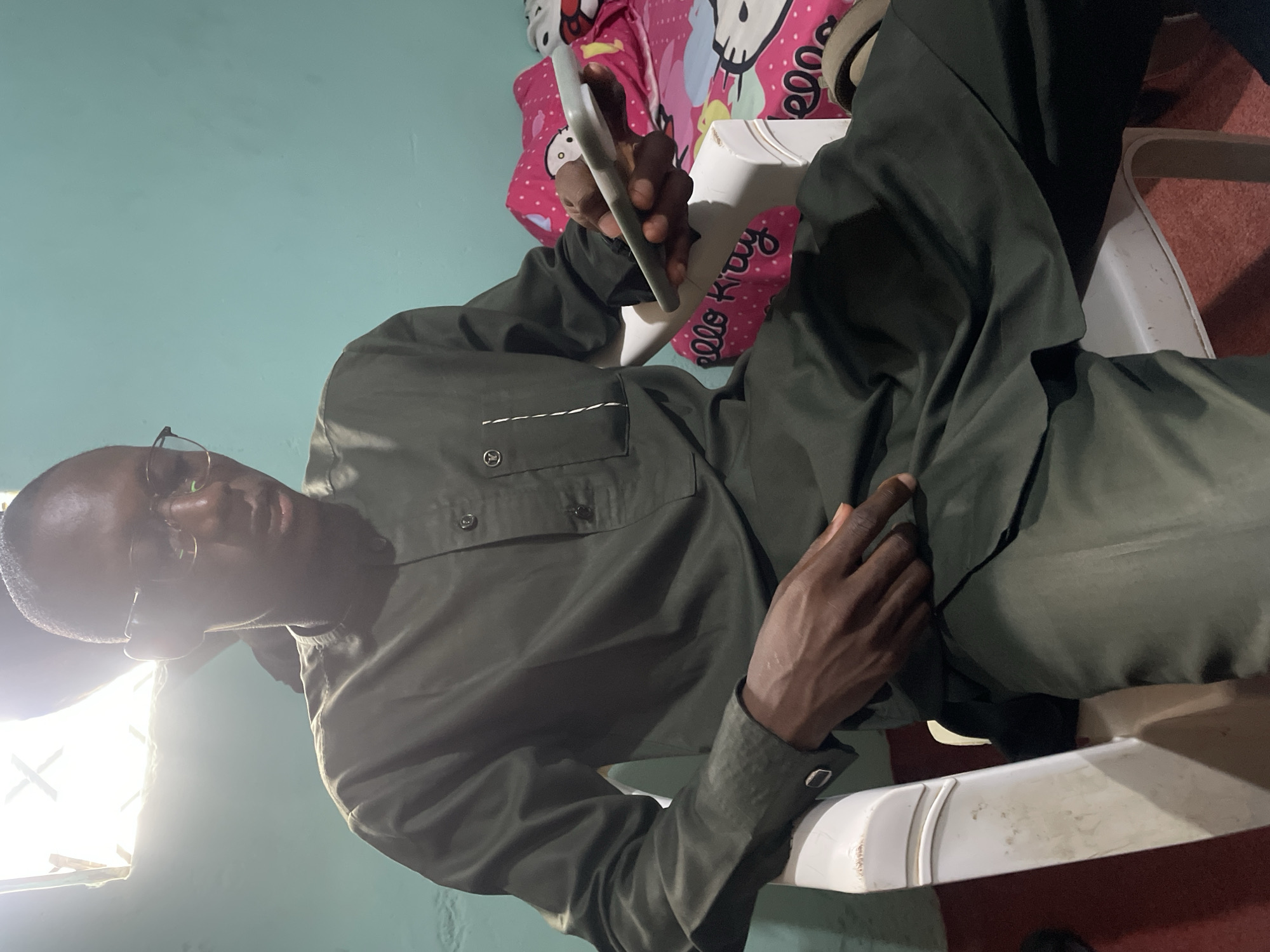 The image shows a man sitting on a white plastic chair. He is wearing glasses and an olive green shirt and pants. He is holding a mobile phone in his right hand. Behind him is a pink and white Hello Kitty pillow. The background shows a light blue wall and a window with bright light. The location is implied to be in the city of Bukuru, Nigeria.