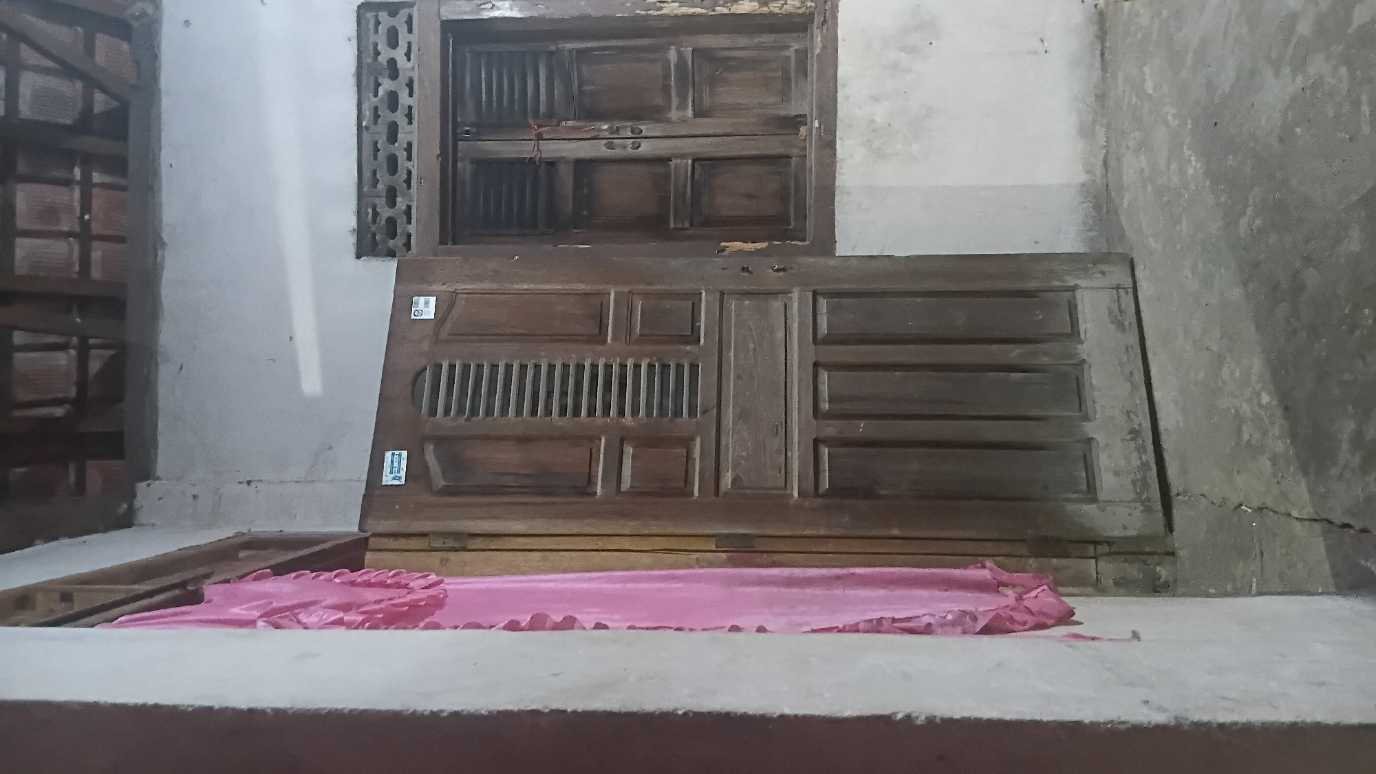 The image shows the exterior of a building in Prabumulih, Indonesia. A brown wooden door with horizontal slats is slightly ajar, revealing a pink curtain behind it. To the right of the door is a wooden window with closed shutters. The walls of the building are rough and unfinished, and the ground is concrete with visible cracks. The roof beams are visible in the upper part of the image. The scene suggests an older, possibly rural or semi-urban dwelling.