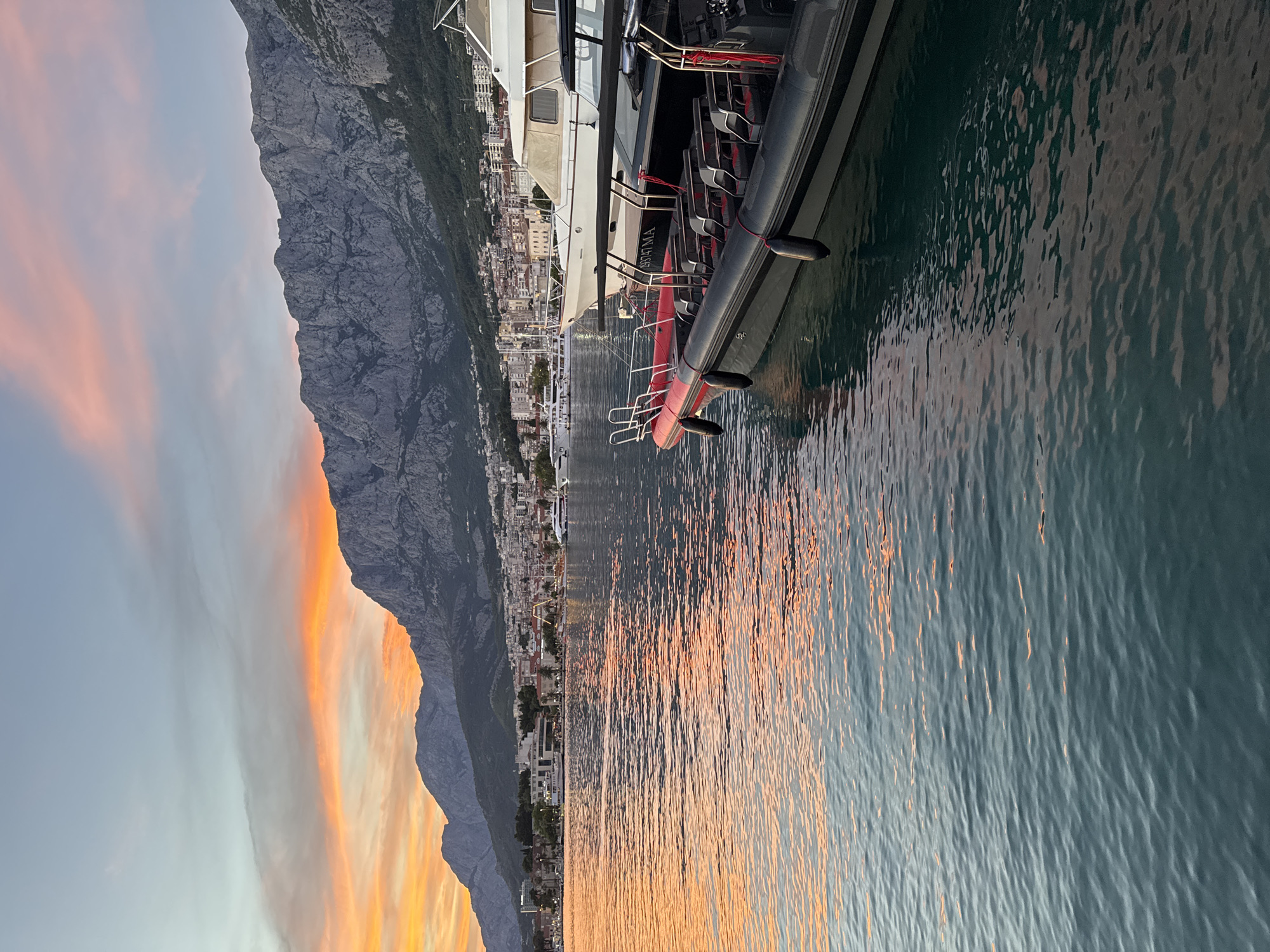 This image captures the scenic Makarska waterfront in Croatia during a sunset. The sky is a canvas of vibrant orange and pink hues, reflecting beautifully on the tranquil water. In the foreground, a black rigid inflatable boat with red accents is docked, its seats neatly arranged. Behind it, a larger white motor yacht is also moored. The backdrop features the city of Makarska, with its buildings nestled at the foot of the imposing Biokovo mountain range, its rugged peaks silhouetted against the colorful sky. The scene is peaceful, with no people visible, emphasizing the natural beauty of the location.