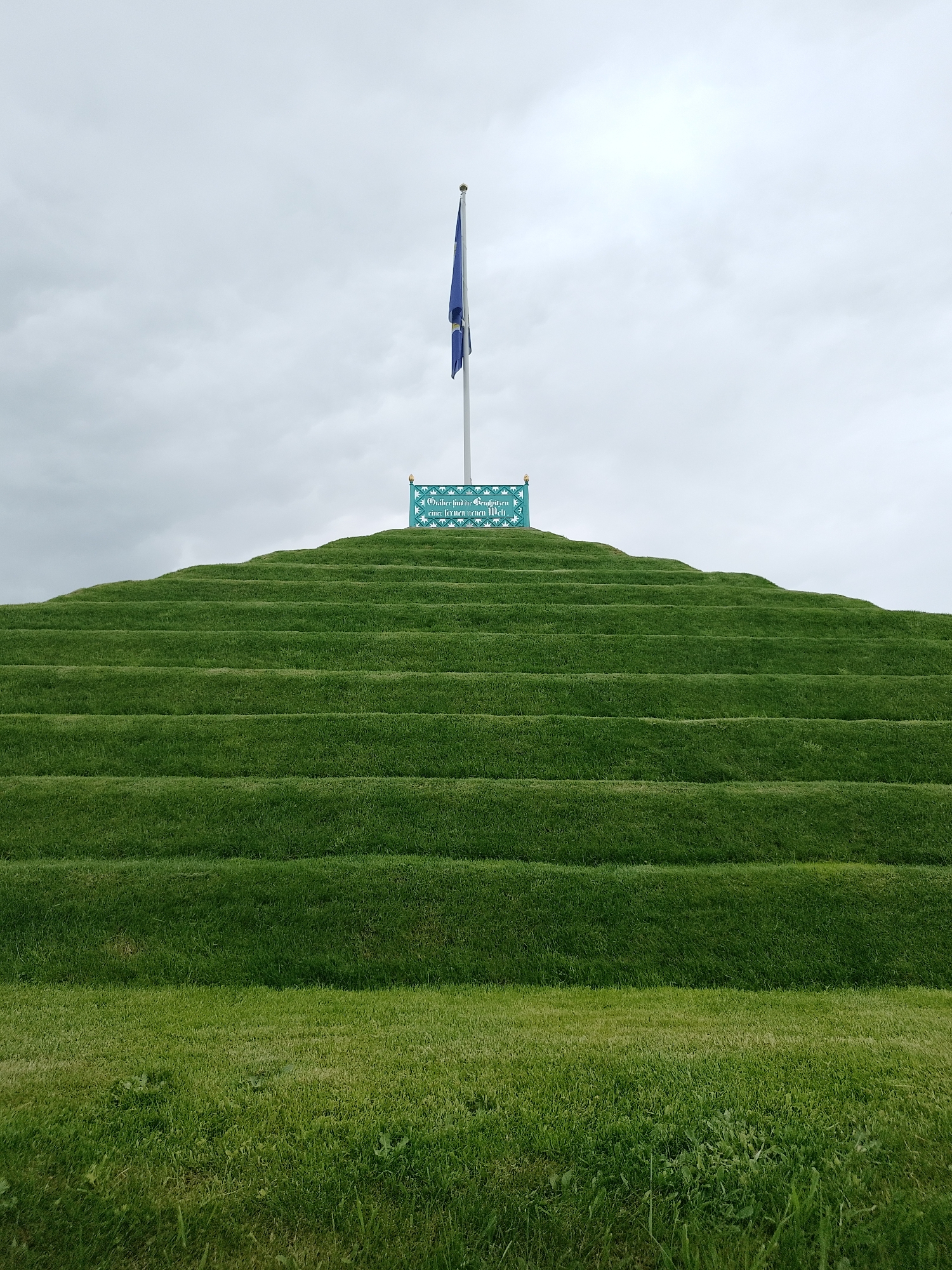 A full shot of the grass-covered pyramidal hill in the city of Magdeburg, Germany, known as the Rotehornpark. The hill consists of numerous stacked layers of grass, resembling steps. A blue flag is placed on top of the hill, along with a rectangular blue sign with white script. The sky is overcast.