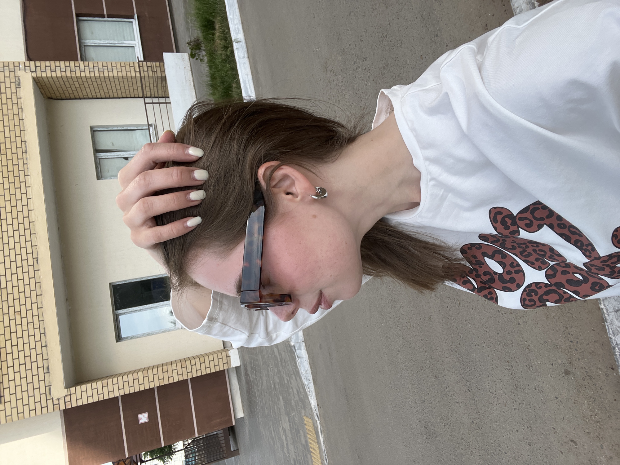 A medium shot, taken from above and angled downwards, presents a woman outdoors during daylight hours. Her right hand is positioned on the back of her head, the fingers subtly splayed. She has fair skin, brunette hair, and an earring is visible in her right ear. Sunglasses are perched on her face. The woman is wearing a white t-shirt adorned with brown, leopard-print-style spots.
In the background, a multi-story building with a light tan brick facade and dark brown accents is discernible. Windows are visible in the building, some with a noticeable reflection. A paved surface occupies the foreground, likely a sidewalk or street. Sparse vegetation is present in the background, suggesting a planted area. The city where it was taken, based on the prompt, is Chaikovsky, Russia.