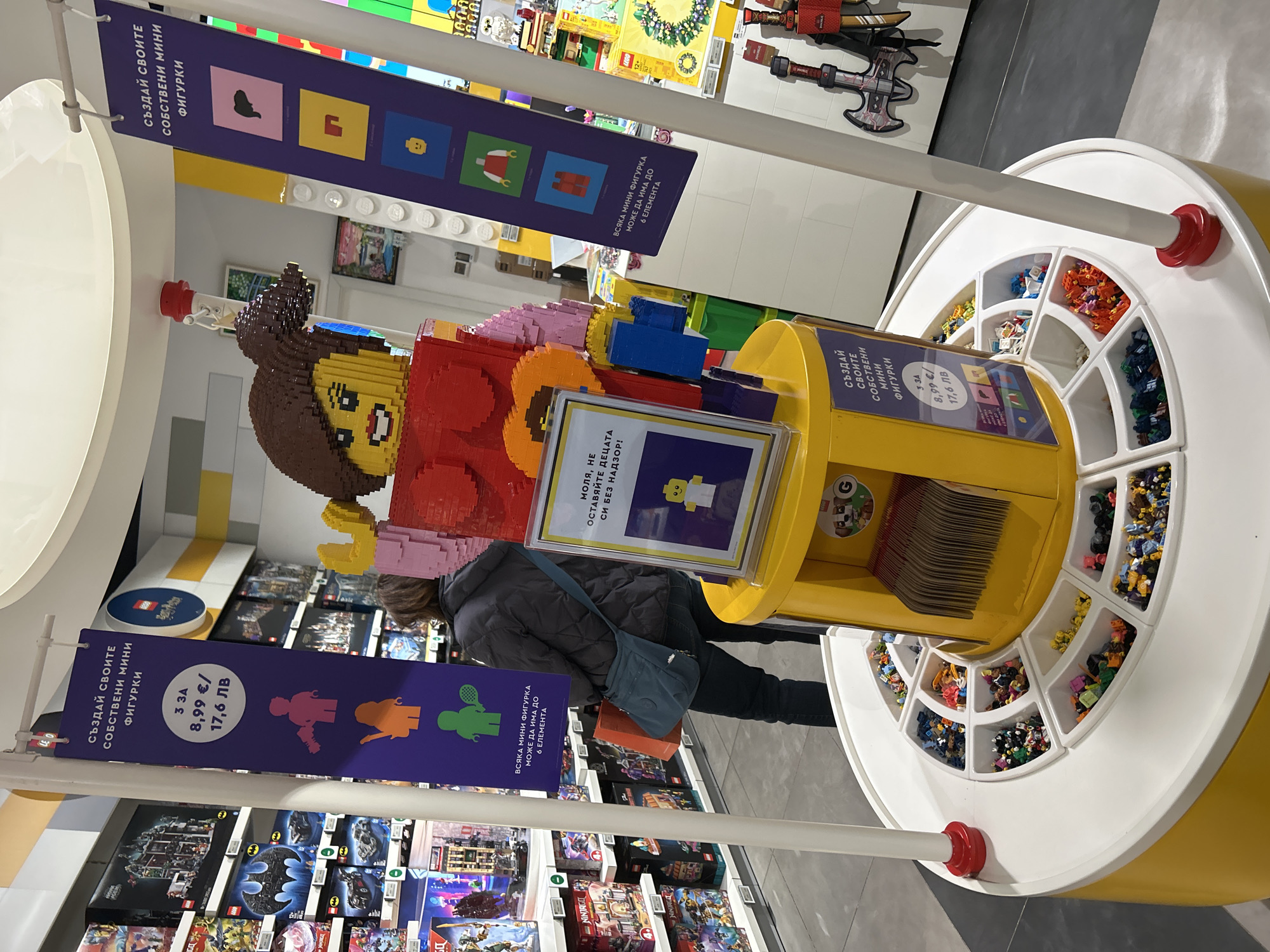 The image depicts an interior scene, likely inside a LEGO store, located in Sofia, Bulgaria. In the foreground, a large LEGO figure of a woman stands next to a "Build a Minifigure" station. The station is cylindrical and yellow with a white countertop that has various compartments filled with LEGO pieces. Signage with the title "Създай своите собствени мини фигурки" (Create your own minifigures) is visible, indicating the purpose of the station, along with price information of "3 3A 8,99 €/ 17,6 ЛВ"
A person wearing a dark jacket and carrying a blue bag is partially visible behind the station. Shelves full of LEGO sets are visible in the background. The overall environment suggests a retail space catering to LEGO enthusiasts.